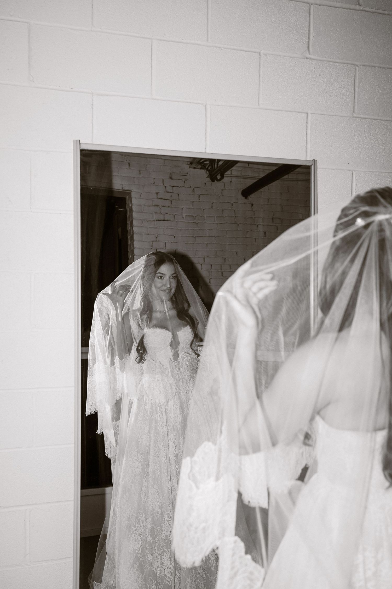 A bride in a white lace wedding gown and veil stands in front of a mirror creating a dreamy reflection.