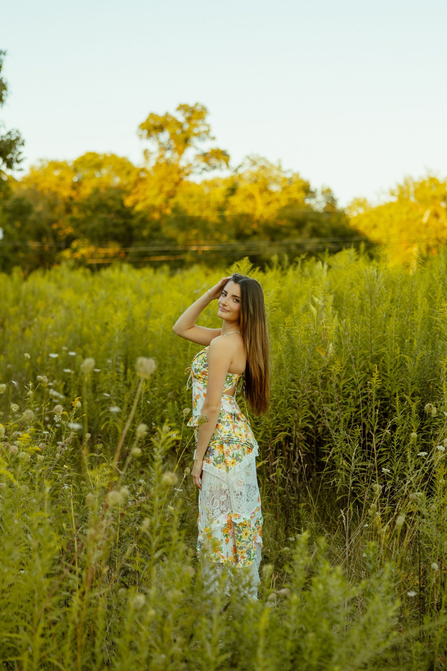 A woman in a floral sundress stands joyfully among tall wildflowers in a golden-lit meadow at sunset.