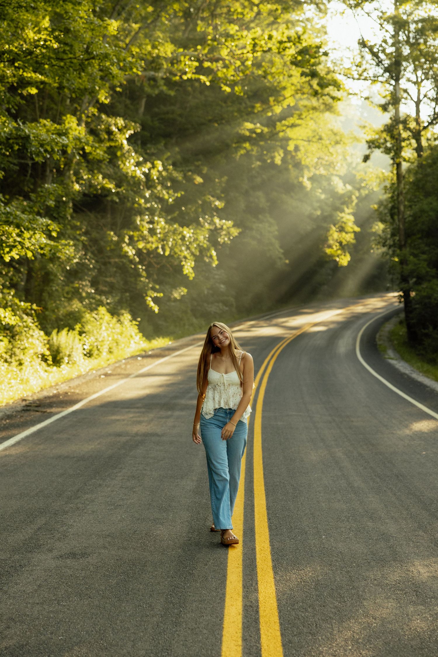 A lone figure walks along a winding forest road as sunbeams filter through green trees on a misty morning.