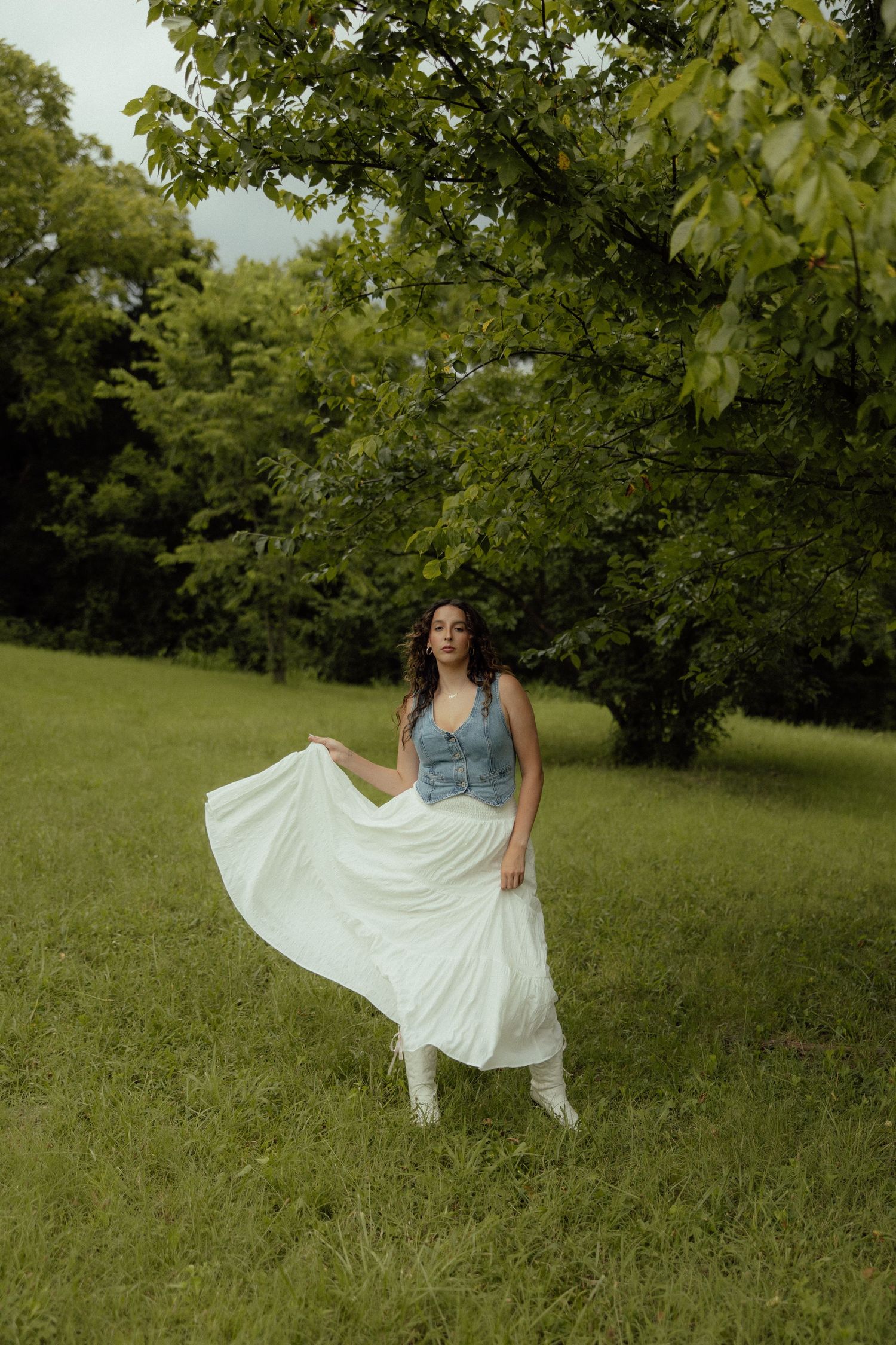 A woman in a white dress twirls gracefully in a sunlit meadow with green trees in the background.