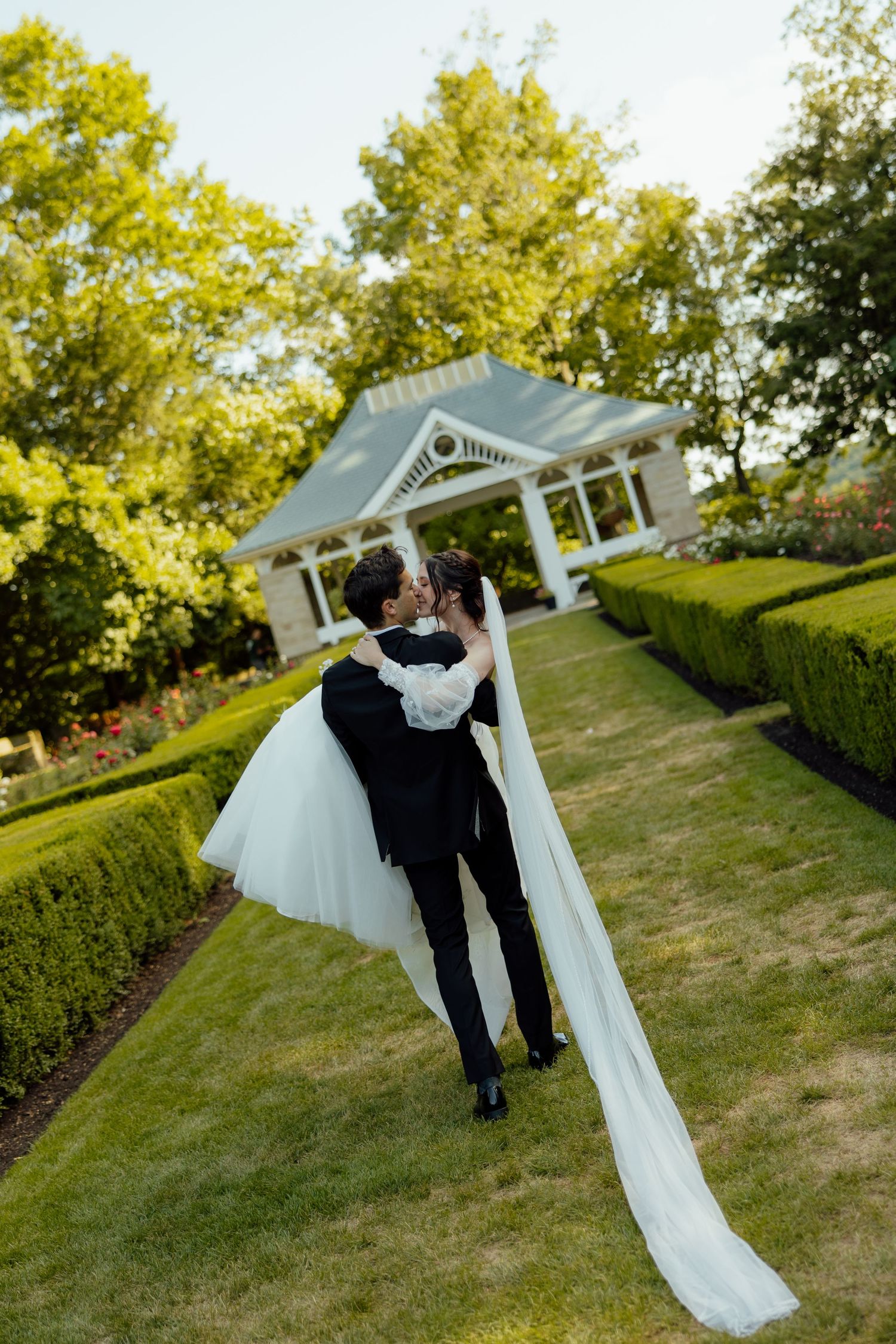 A couple shares a romantic moment under a white garden arbor surrounded by lush greenery on a sunny day.