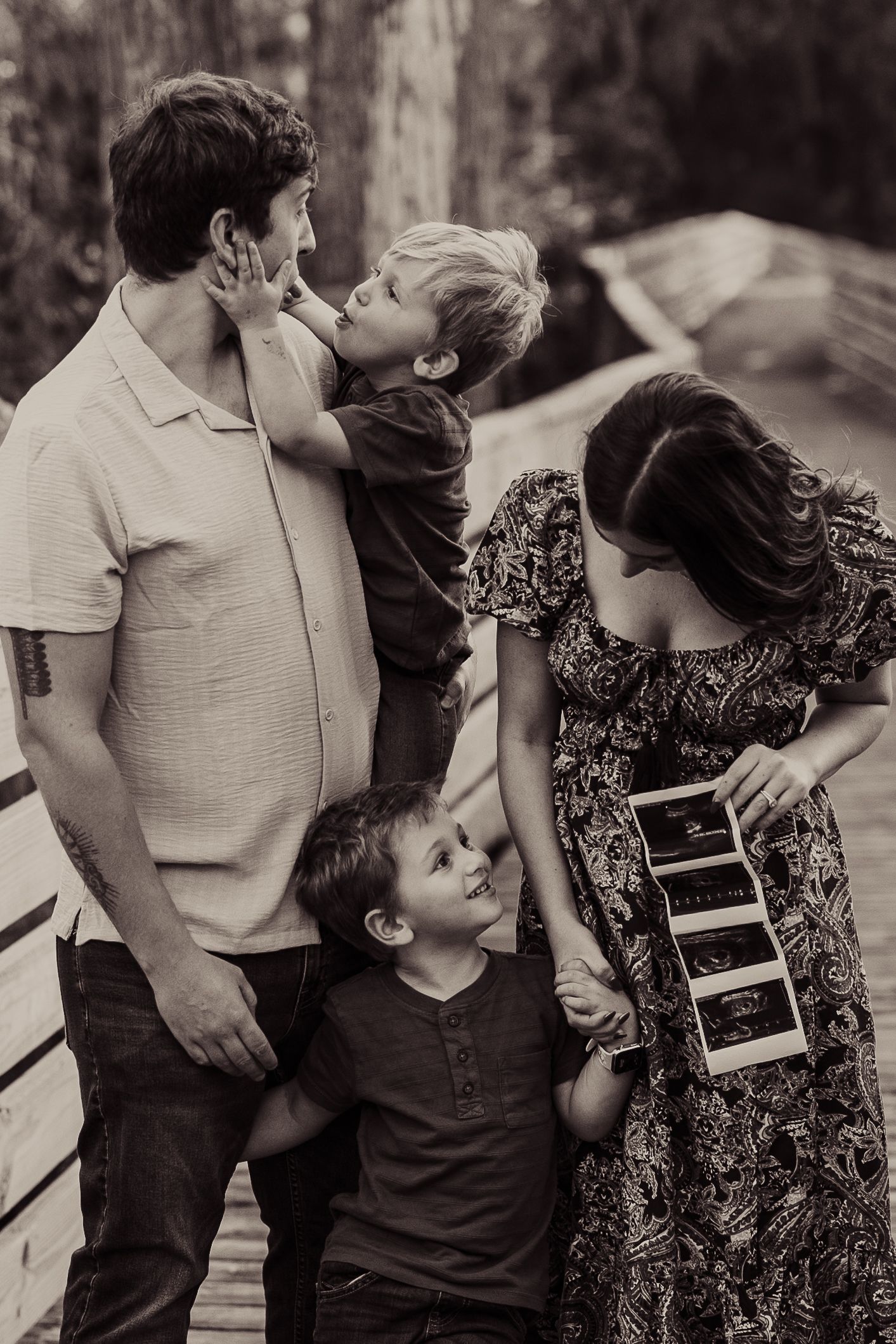 Family gathered on wooden deck looking at ultrasound photos together in black and white.
