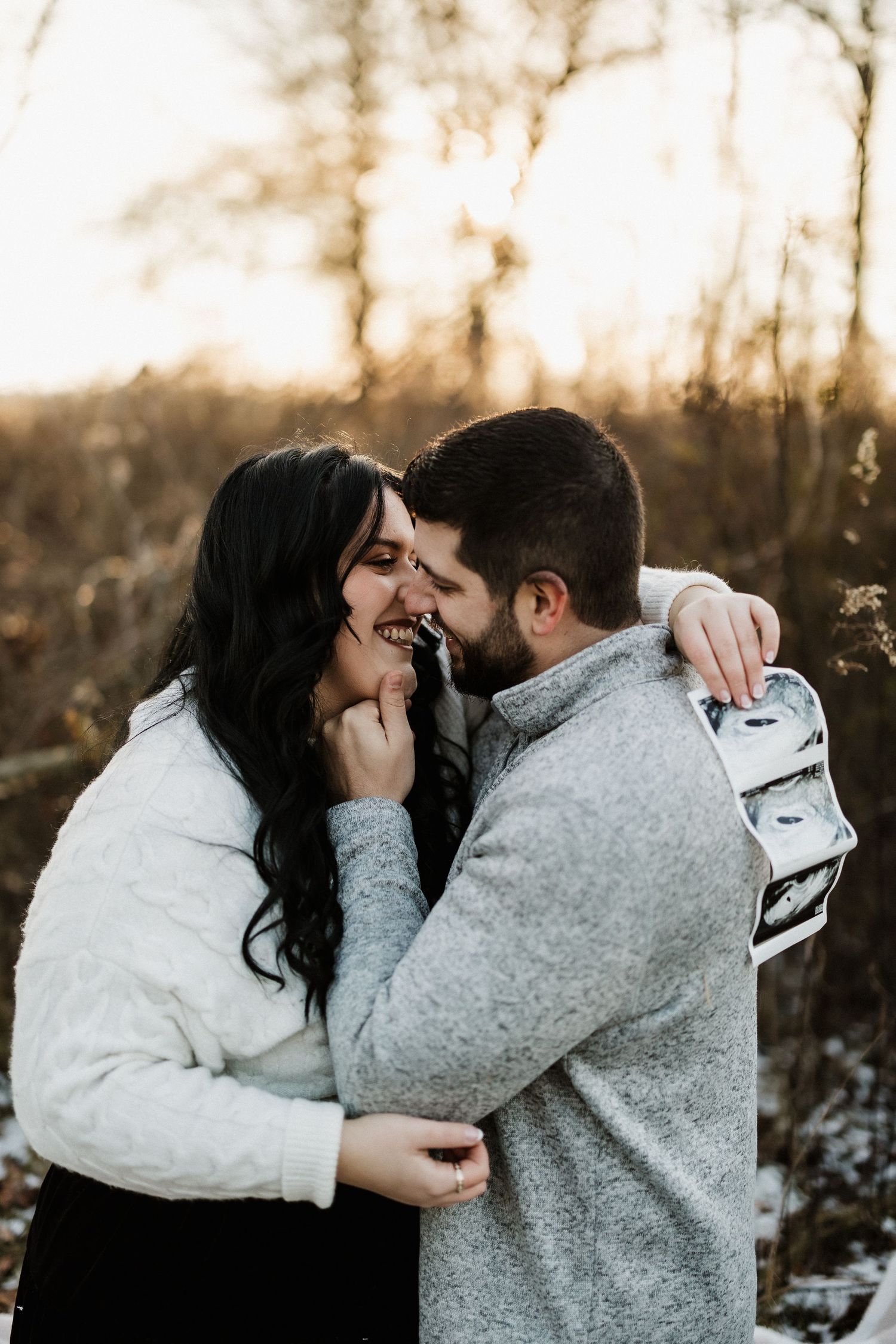 Couple embracing outdoors while holding ultrasound photos during sunset.