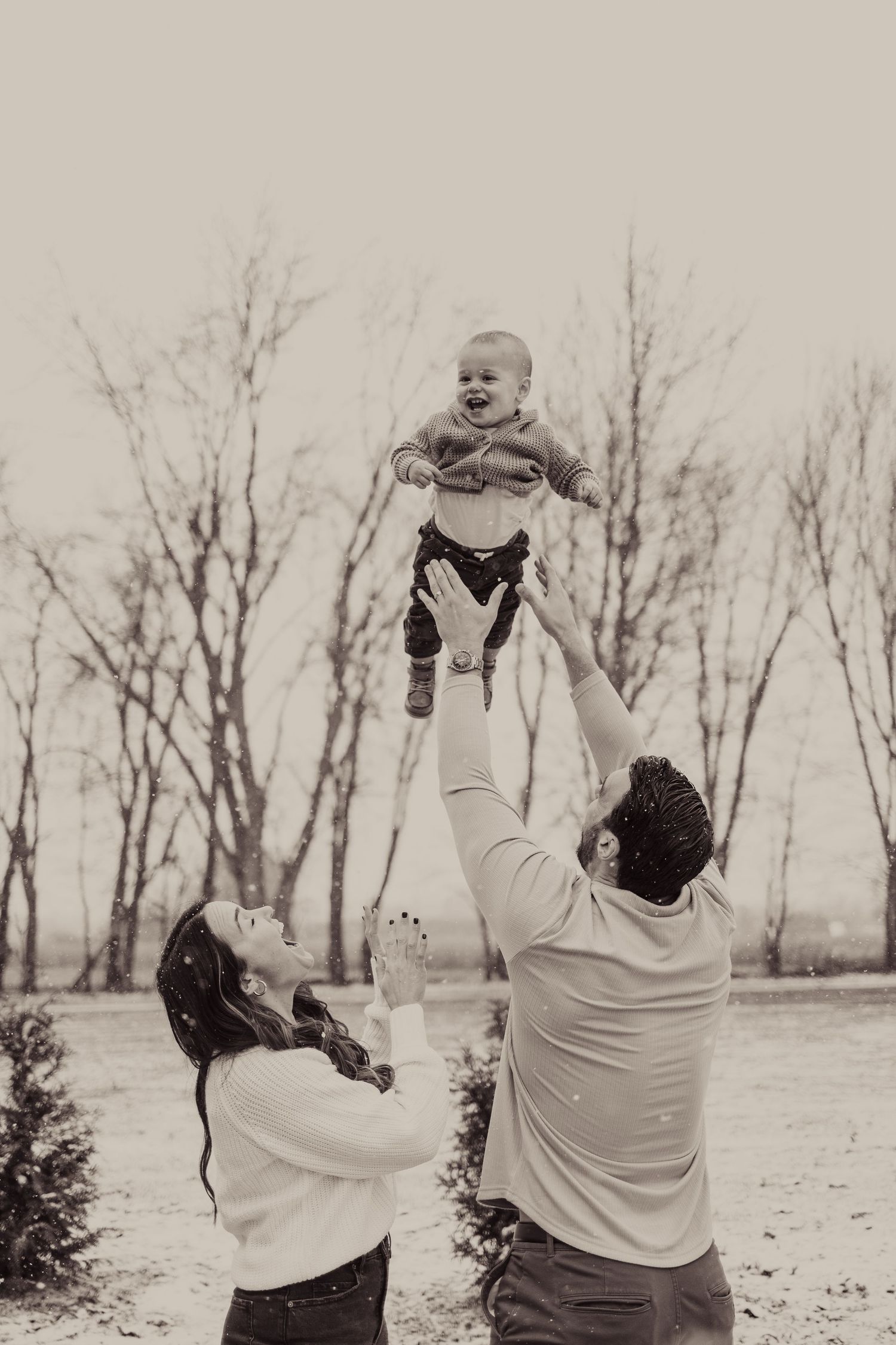 Parents playfully lift small child in the air against winter landscape.