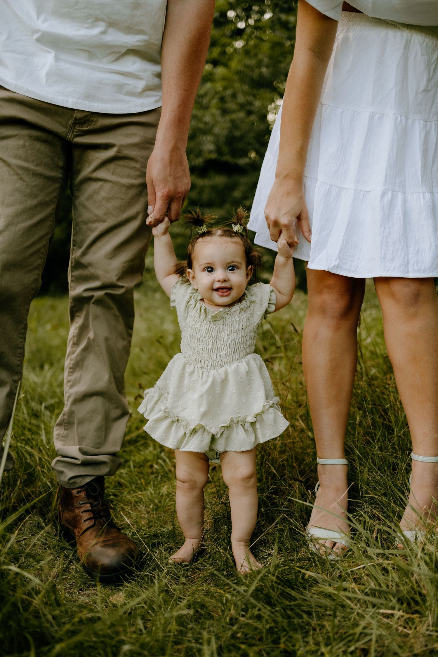 Baby in white dress standing between parents' legs in grassy field.