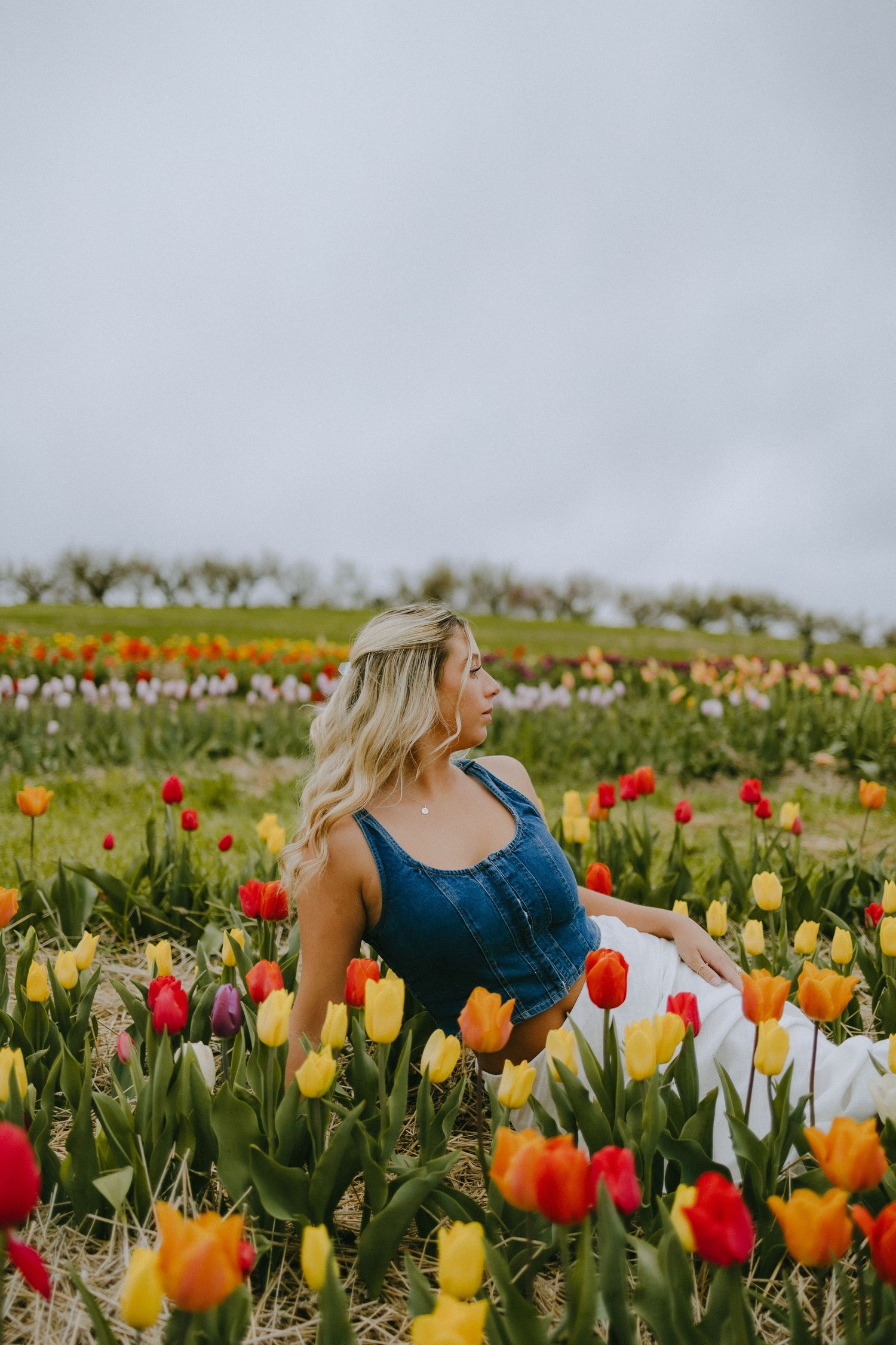Woman in blue dress sitting among vibrant yellow, red and orange tulips on a cloudy spring day.