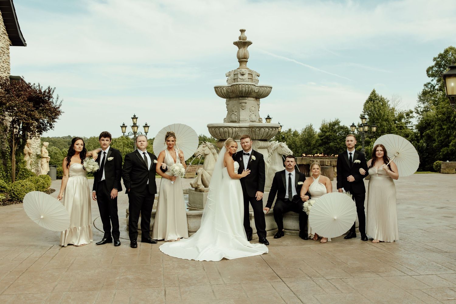 Wedding party poses outdoors in formal attire with parasols and bouquets on a sunny day.