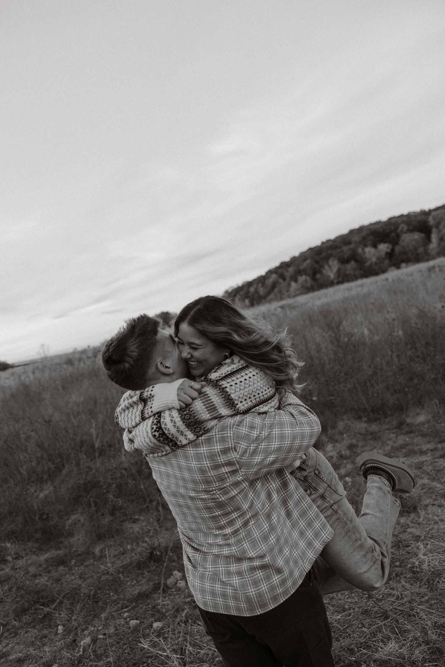 Black and white photo of two people embracing on a grassy hillside with dramatic cloudy sky in the background.
