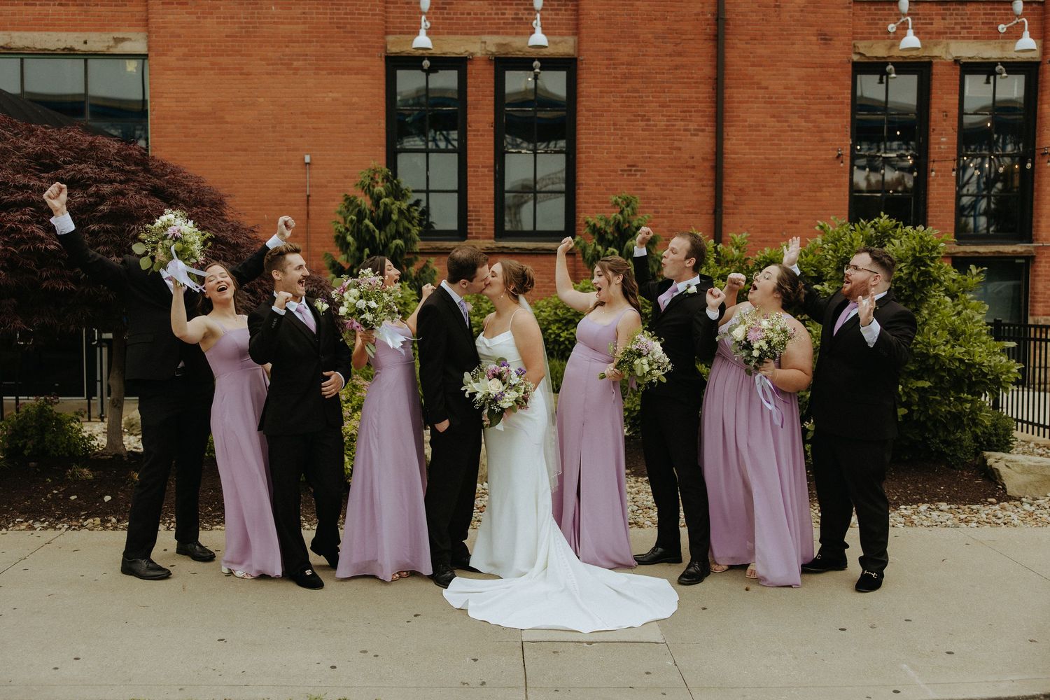 Wedding party in purple bridesmaid dresses poses outdoors against brick building backdrop.