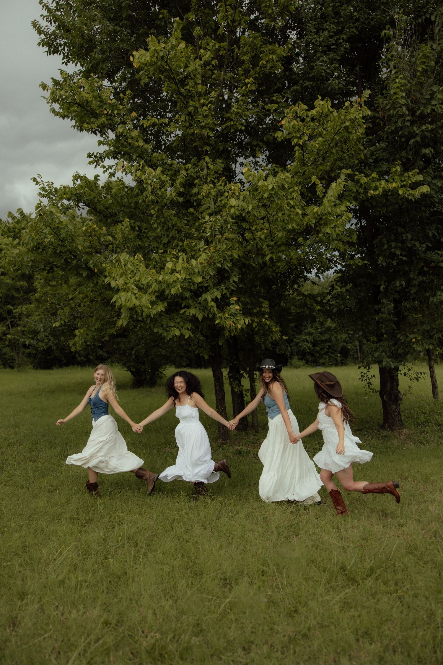 A group in white dresses lies playfully on a grassy hillside beneath trees on a cloudy day.