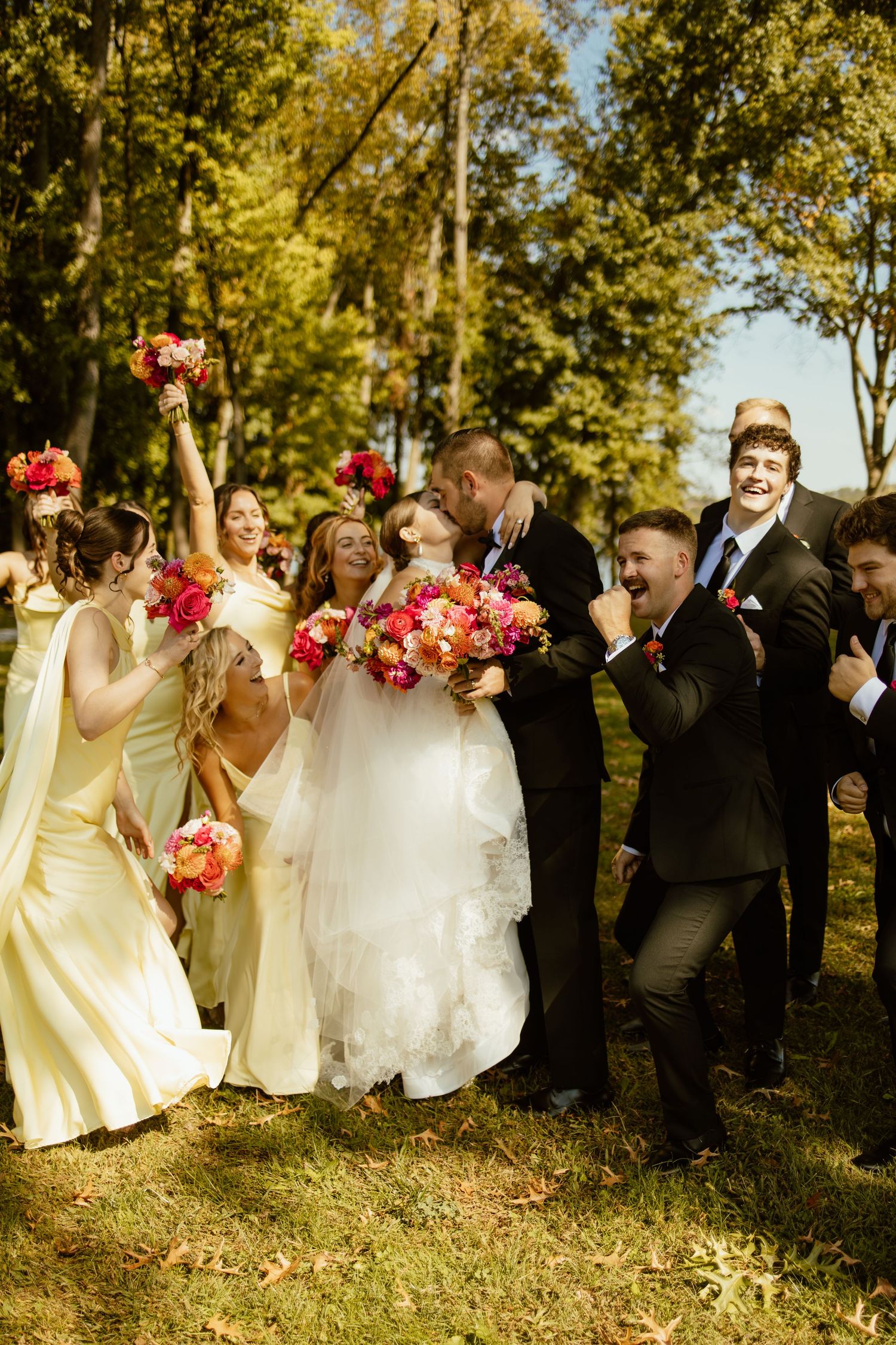 Wedding party stands in a line against autumn trees with yellow and pink floral bouquets.