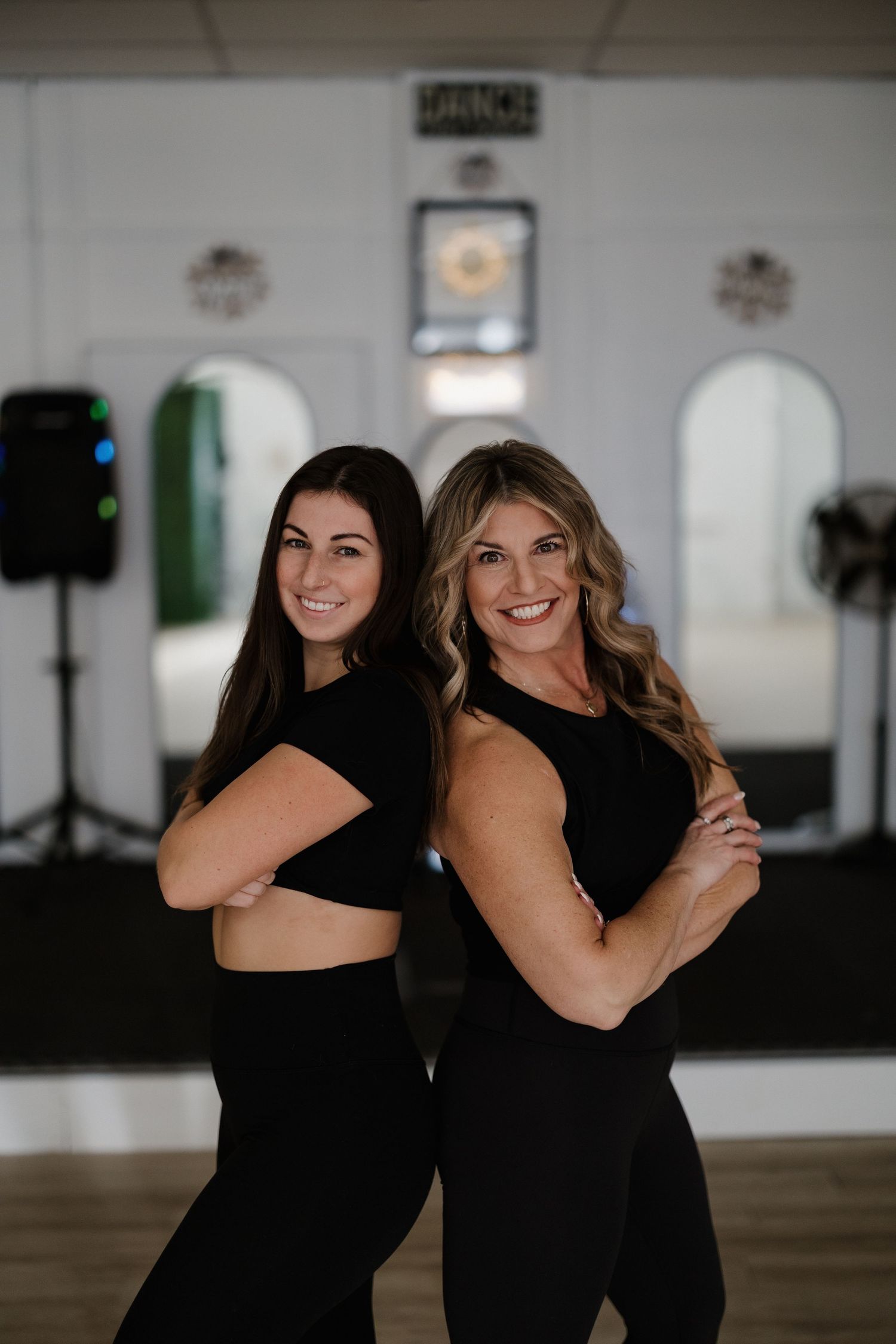 Two fitness instructors pose back-to-back in matching black athletic wear in a dance studio.