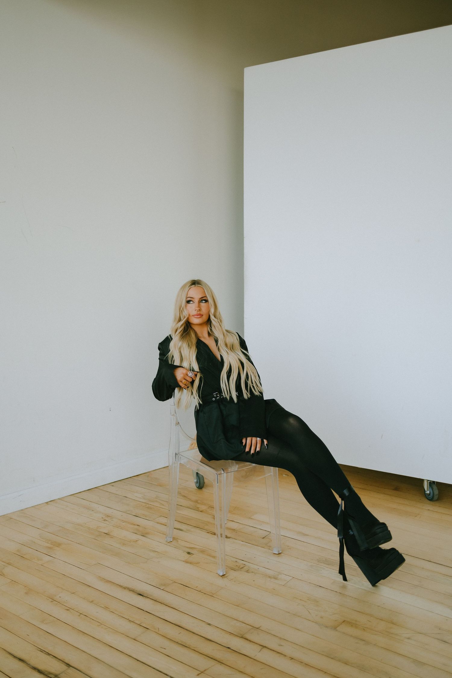 Fashion portrait in black ensemble seated on clear chair against minimal background.
