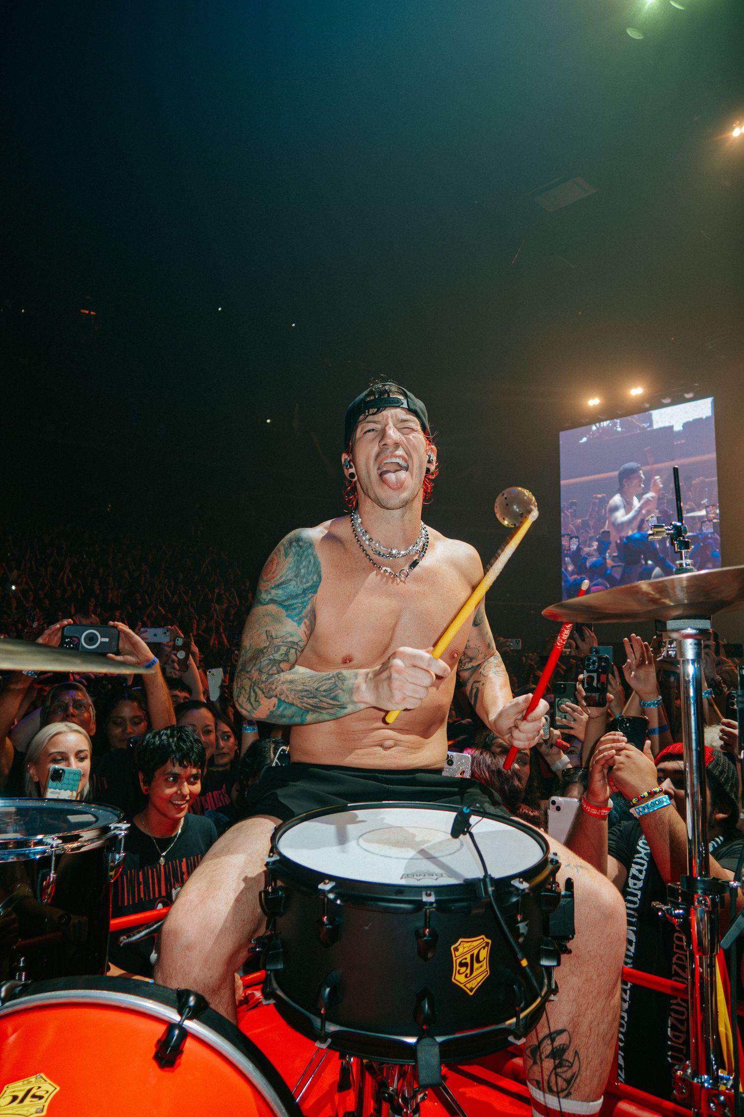 Shirtless drummer with tattoos performs energetically on stage during a concert while crowd cheers.