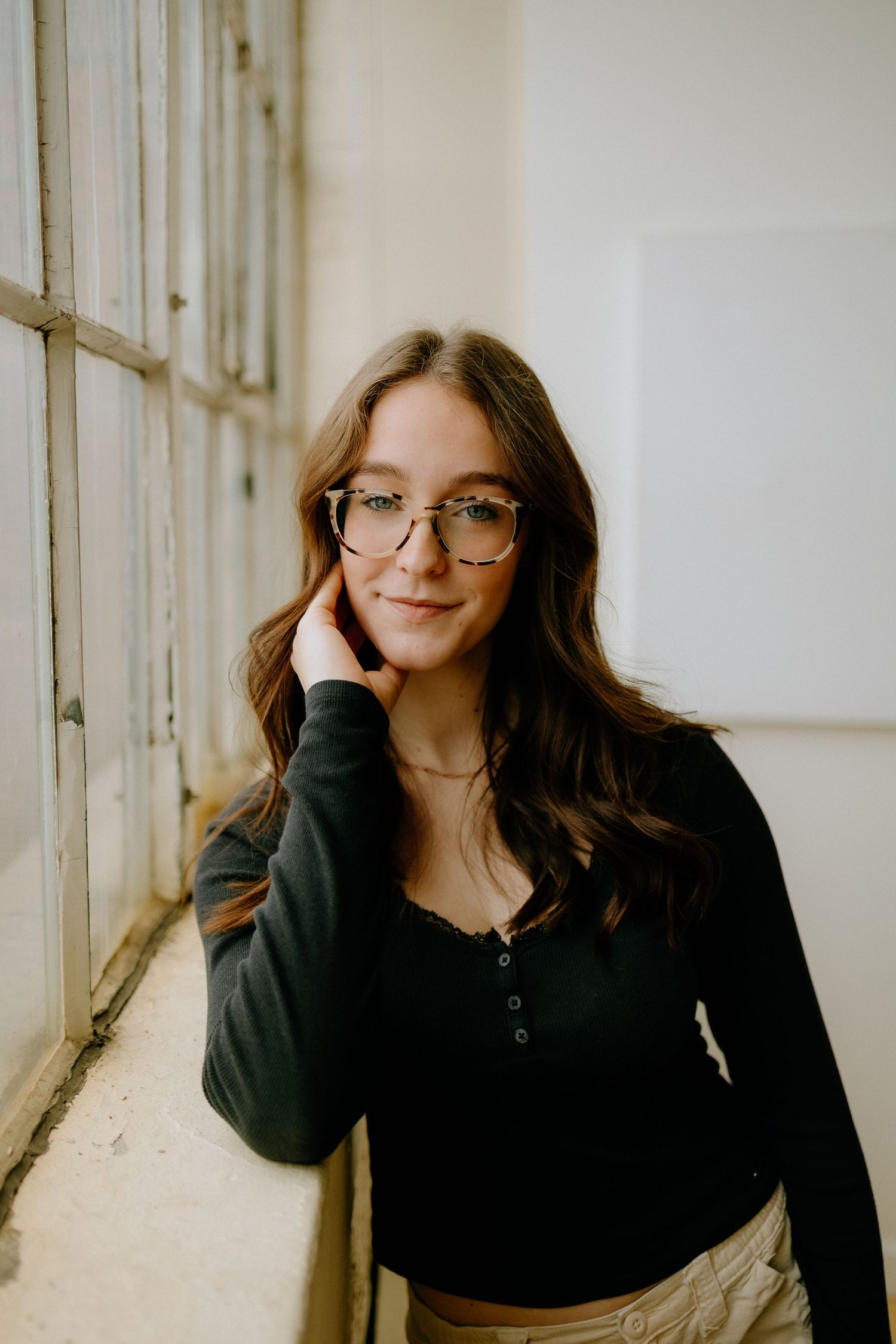 A portrait of someone in glasses and black sweater posing by a window in natural light.