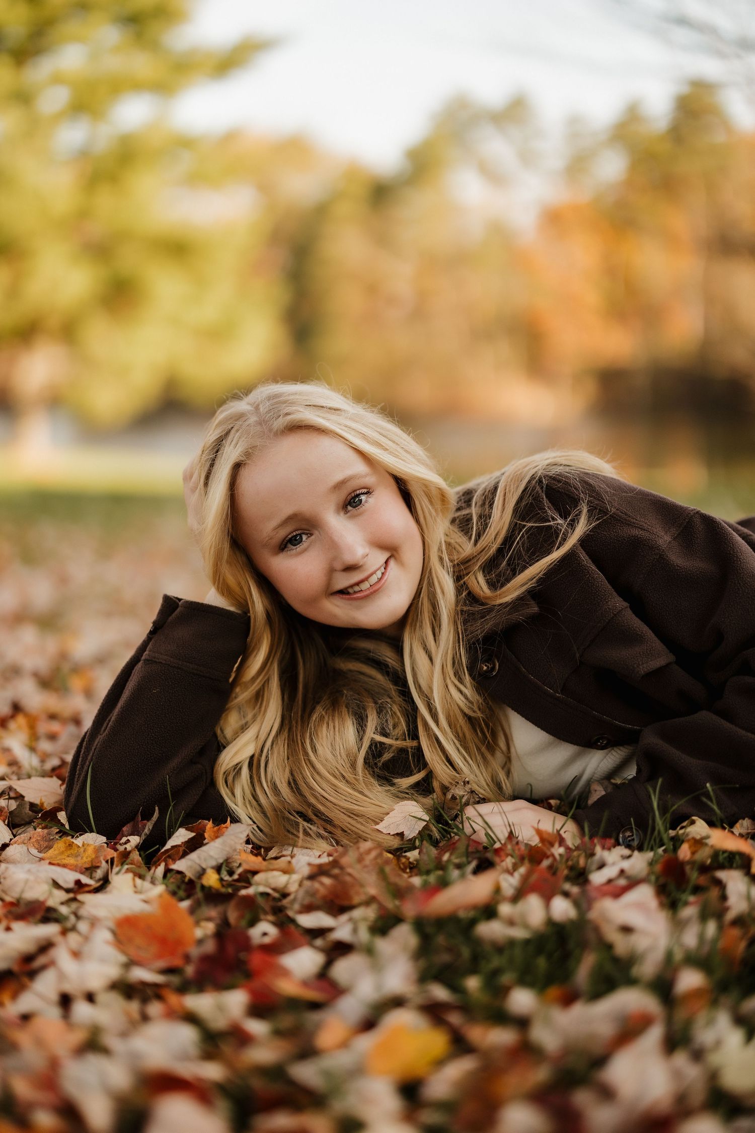 A candid autumn portrait of someone laying in colorful fallen leaves wearing a brown jacket.