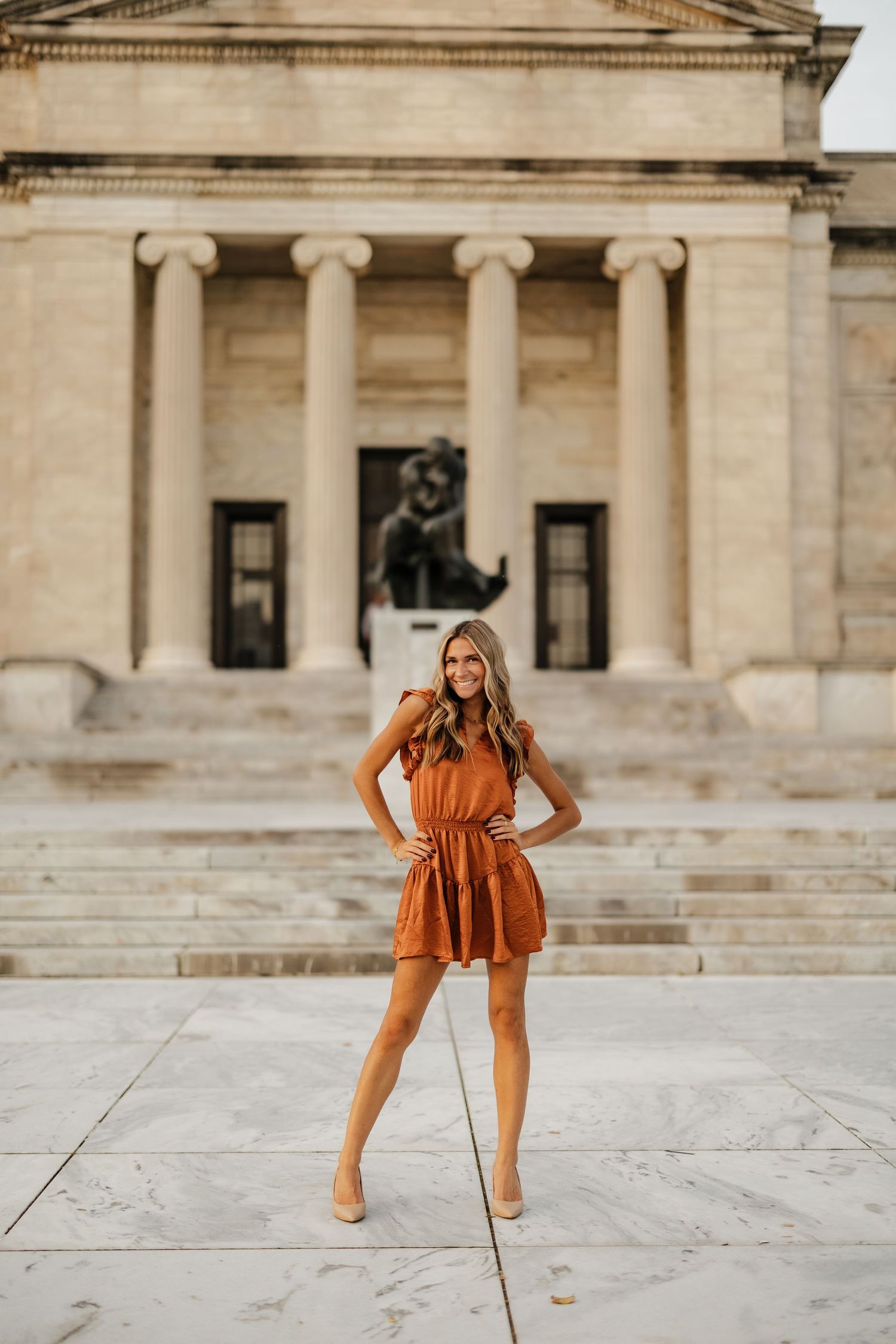 Someone in a rust-colored dress poses confidently in front of a neoclassical building with columns.