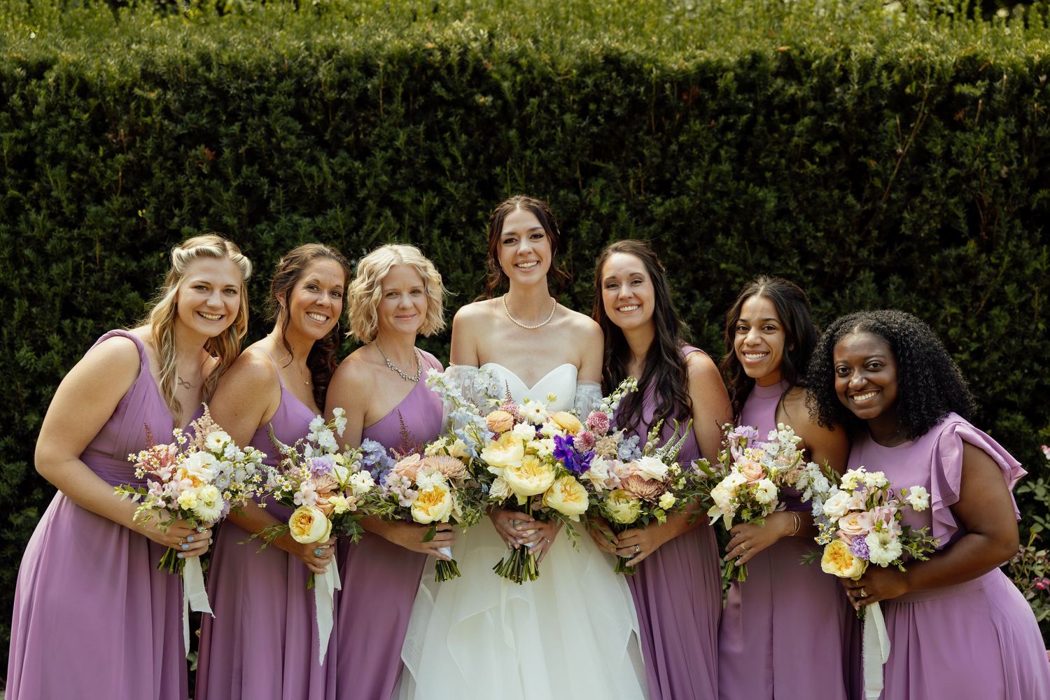Bridesmaids in lavender dresses holding colorful bouquets pose together in garden setting.