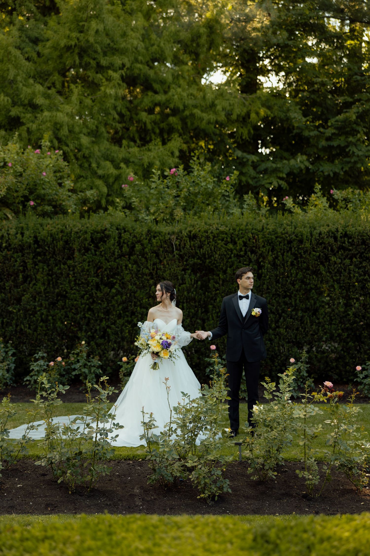 A wedding couple shares an intimate moment in a beautiful garden setting with tall hedges and blooming flowers.