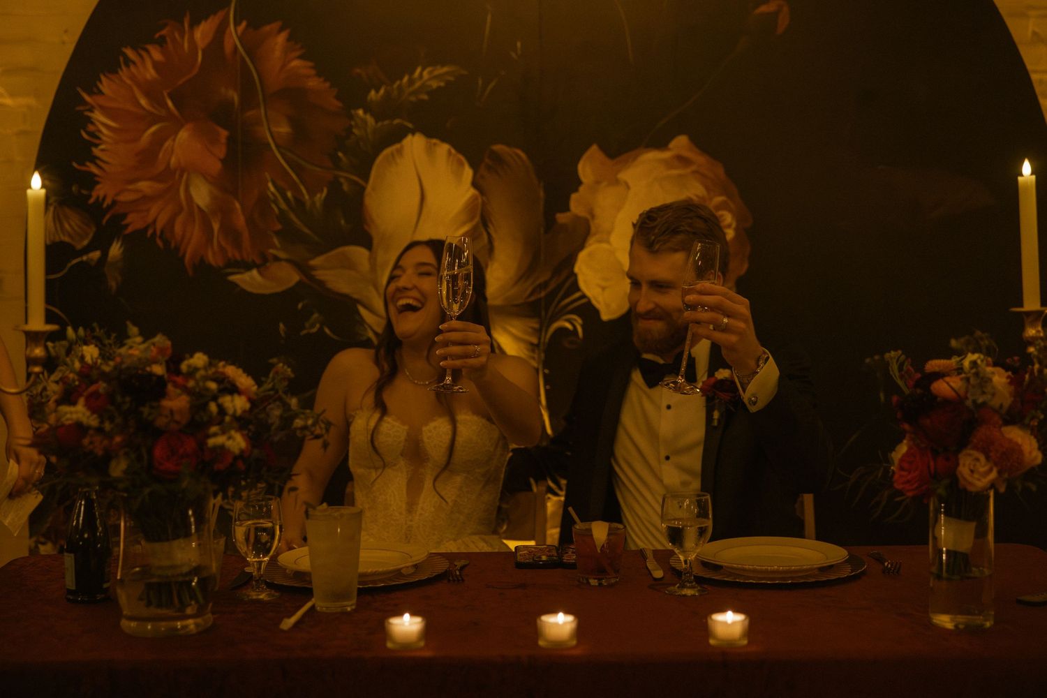 Two people take photos with cameras at a dimly lit table decorated with flowers and candles.