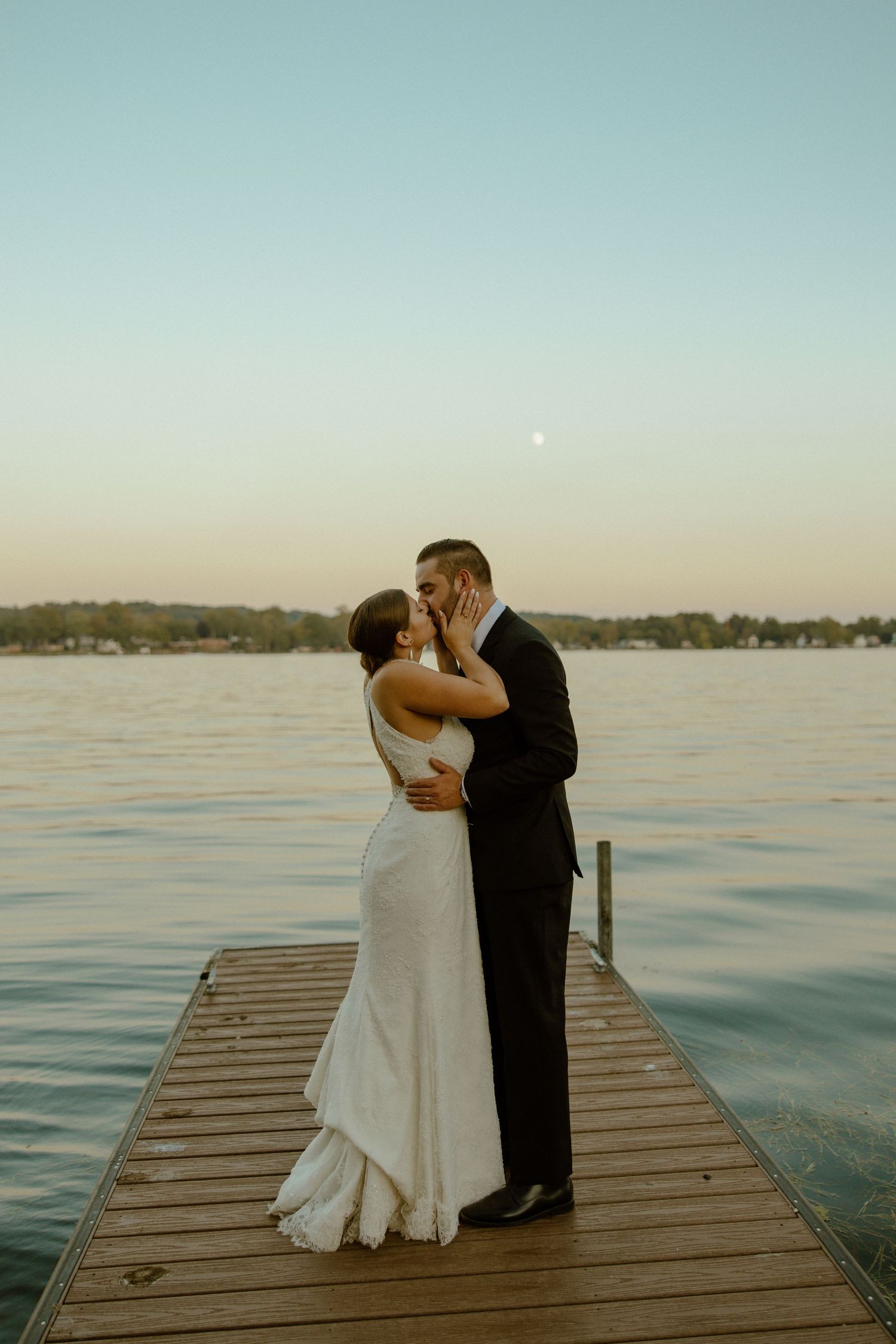 A romantic couple shares a tender kiss on a wooden dock at sunset over a tranquil lake.