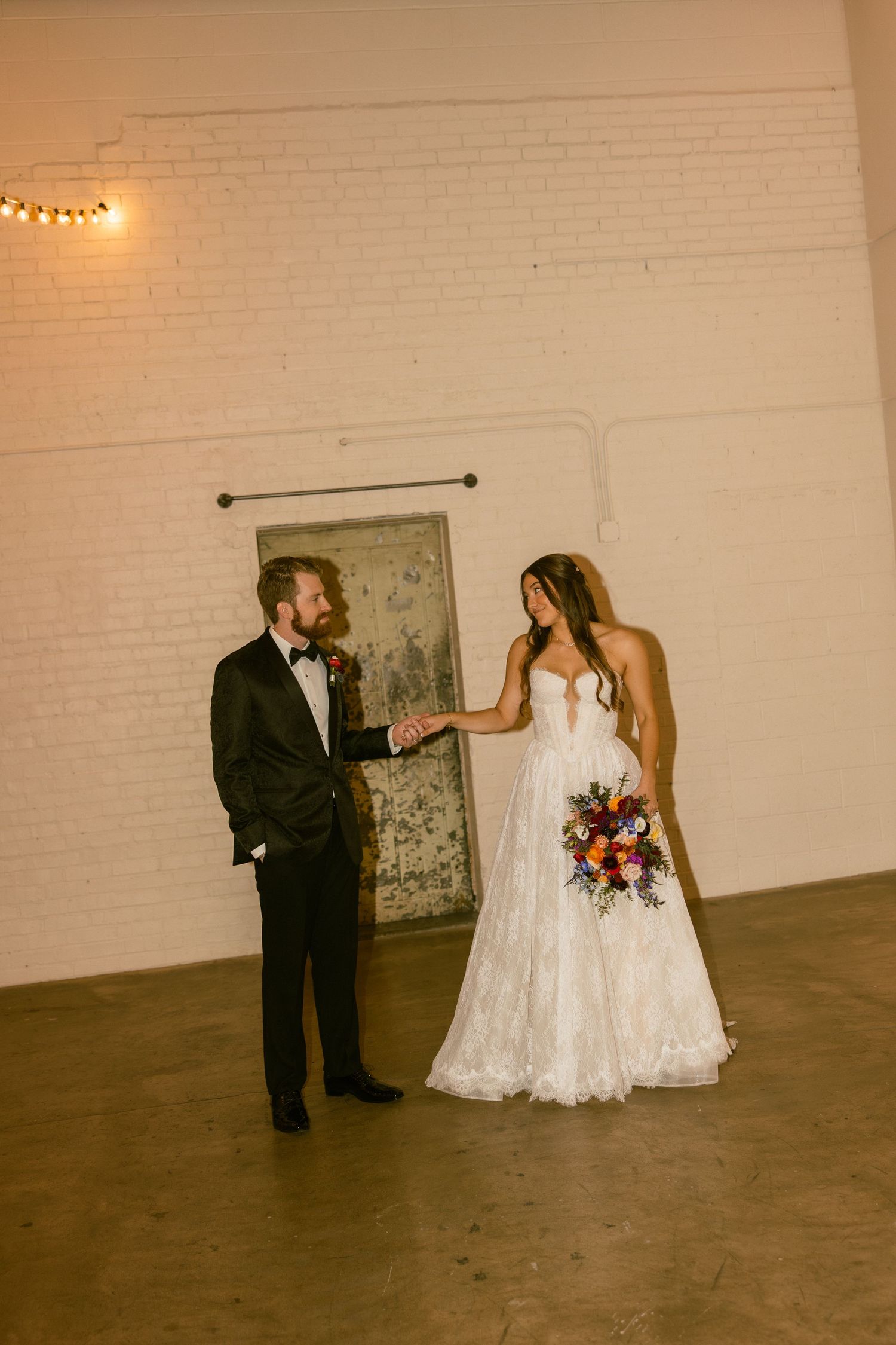 A wedding couple dances together in a minimalist industrial space with white brick walls and string lights overhead.
