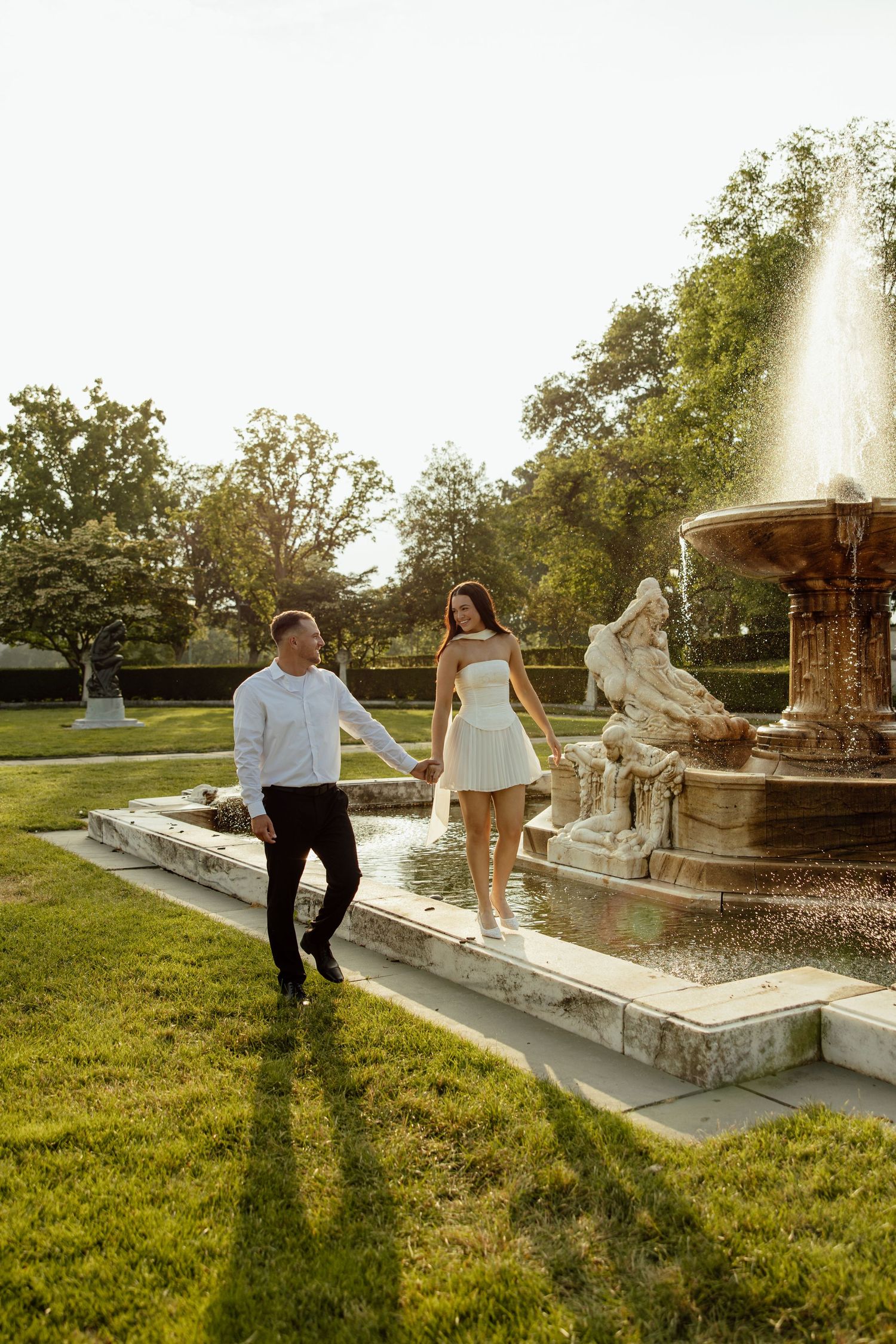 A couple shares a playful moment by an ornate fountain in a sunlit garden during a photo session.