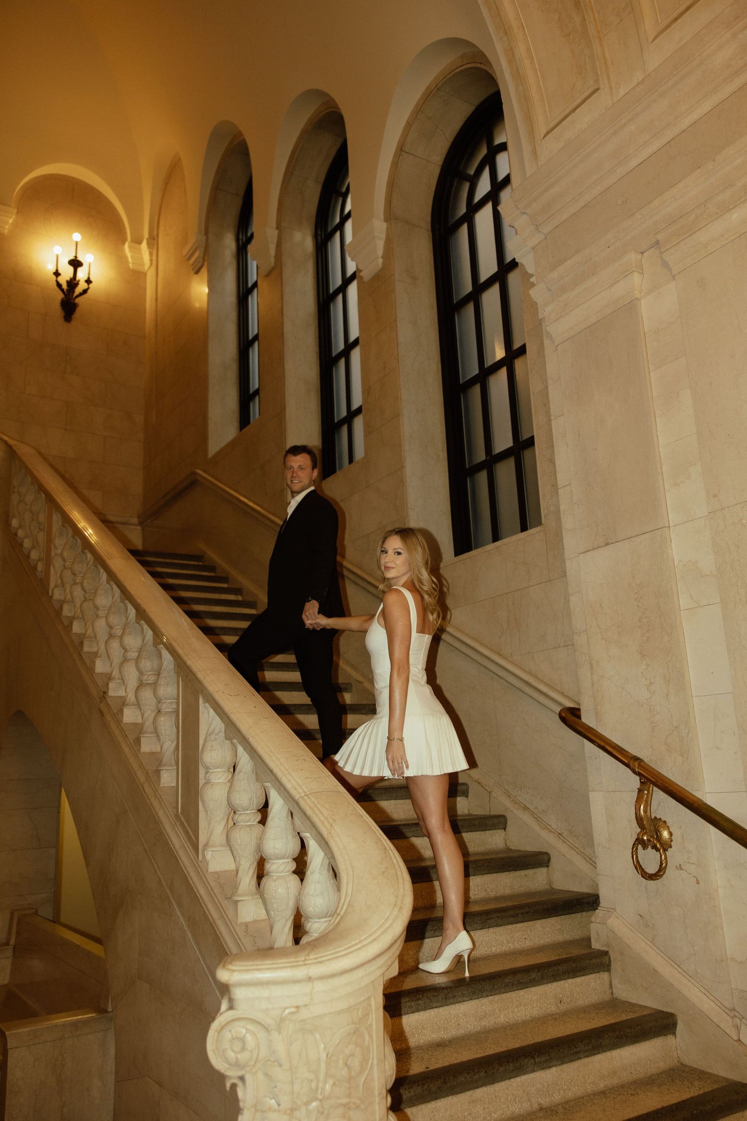 A person in a white dress poses on an elegant marble staircase in a historic building interior.