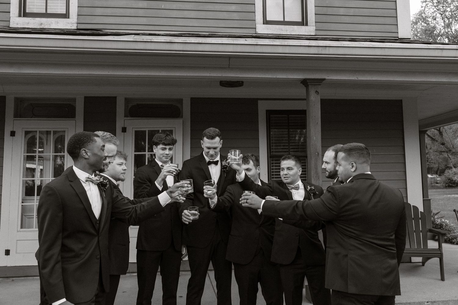 Groomsmen in black suits and bowties stand in line on a covered porch of a gray house.