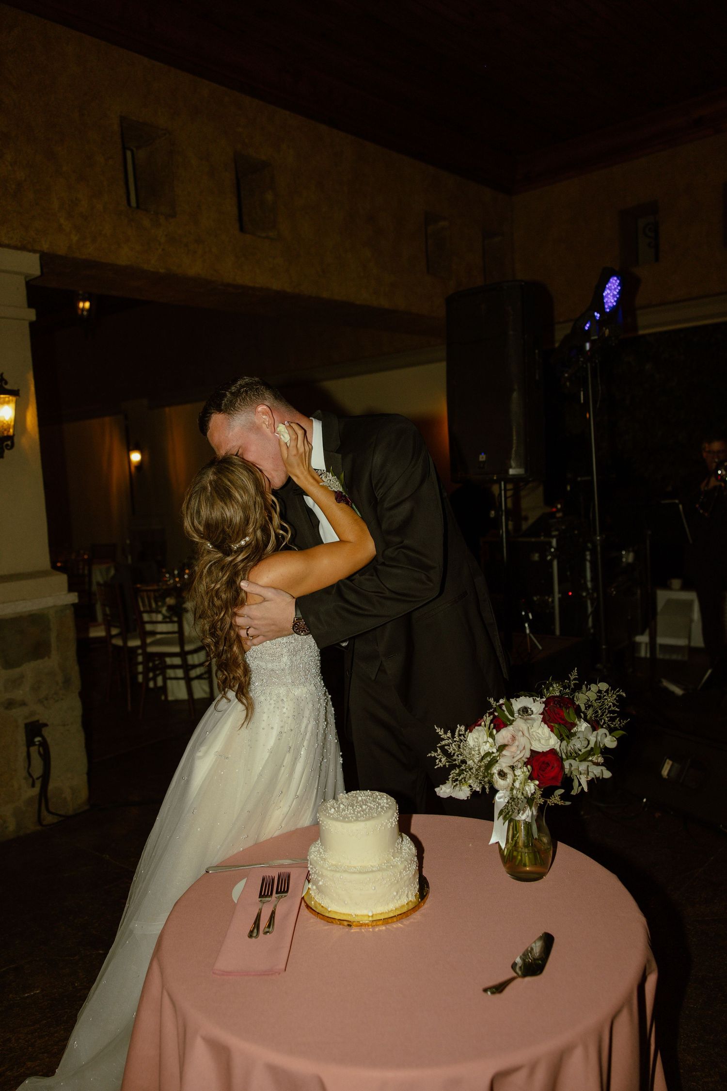 Wedding reception setup features a pink tablecloth with white cake and floral centerpieces in dim lighting.