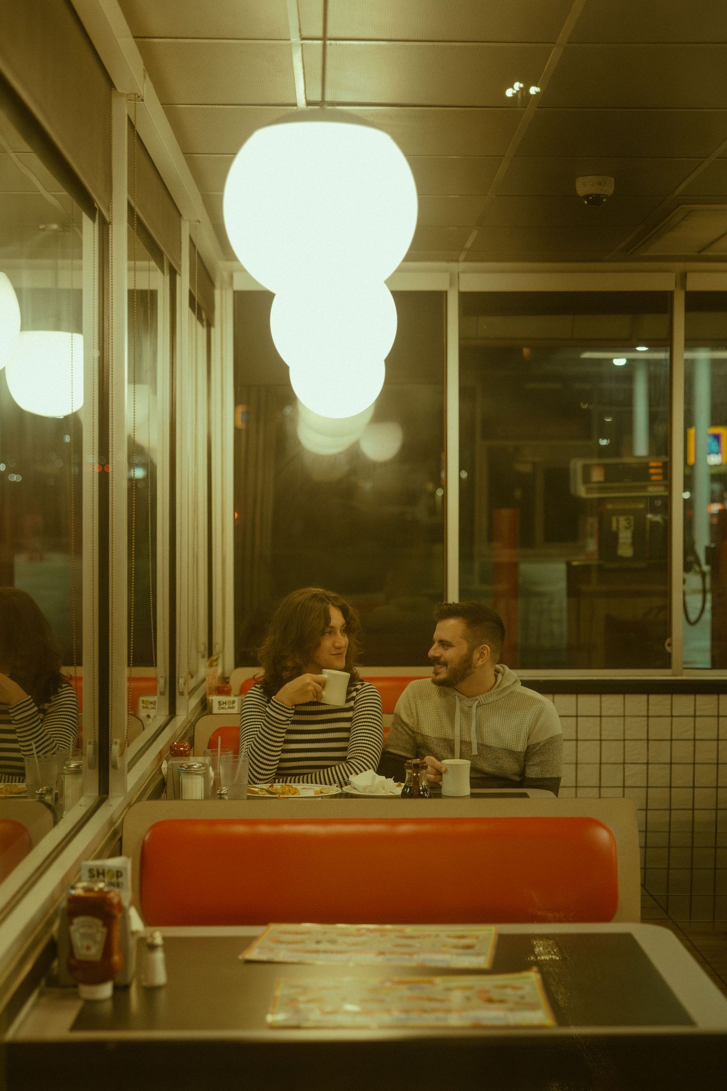 Diners seated in retro-style restaurant booths with glowing round pendant lights creating moody evening ambiance.