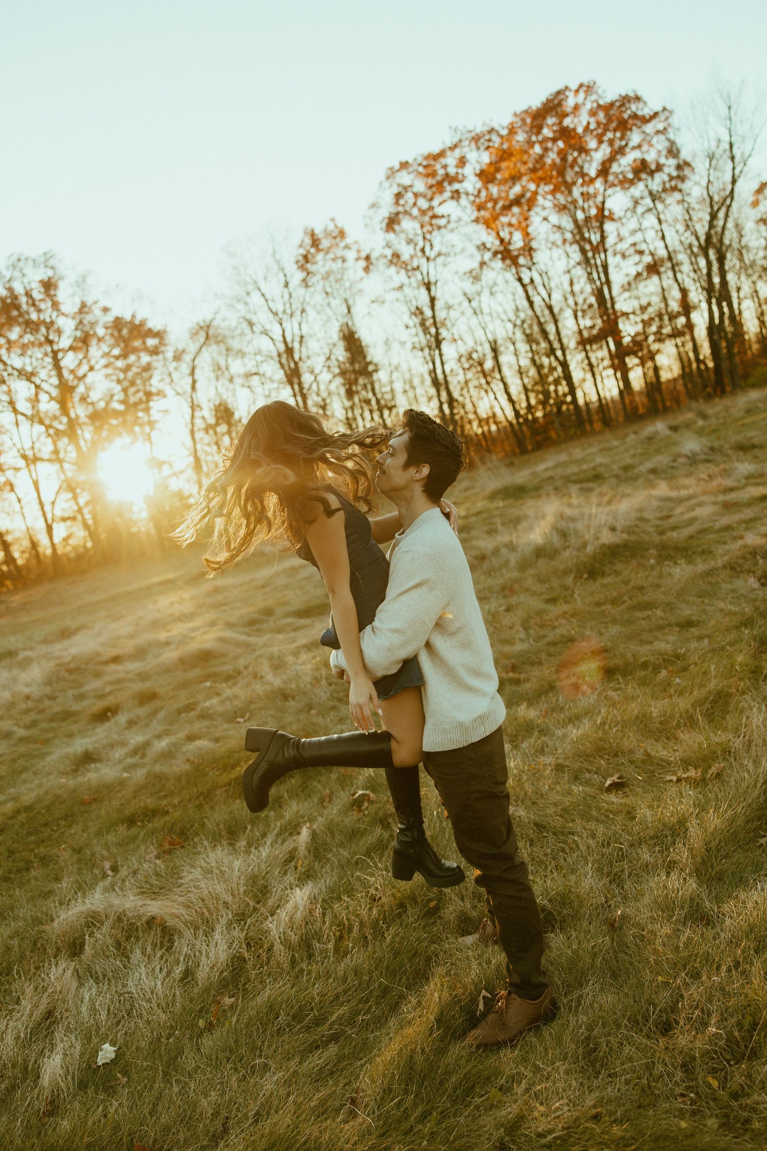 A couple embraces in a joyful dance pose on a grassy hillside during golden hour sunset with autumn trees in background.