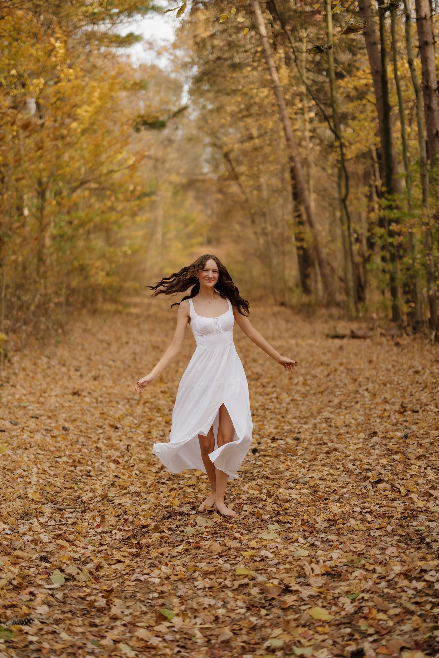 A flowing white dress twirls through fallen leaves on a tree-lined autumn path.