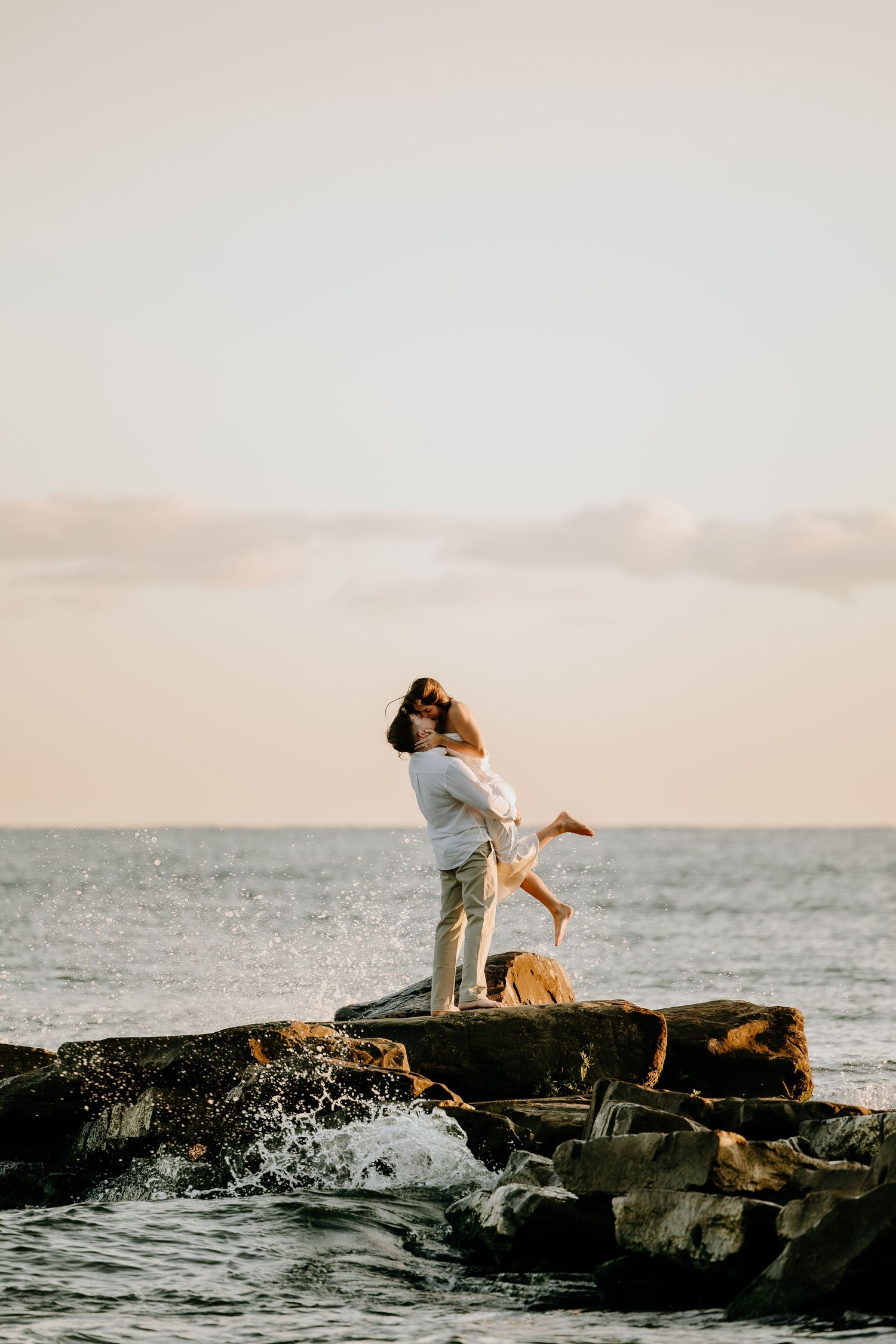 A couple celebrates on coastal rocks as waves crash below.