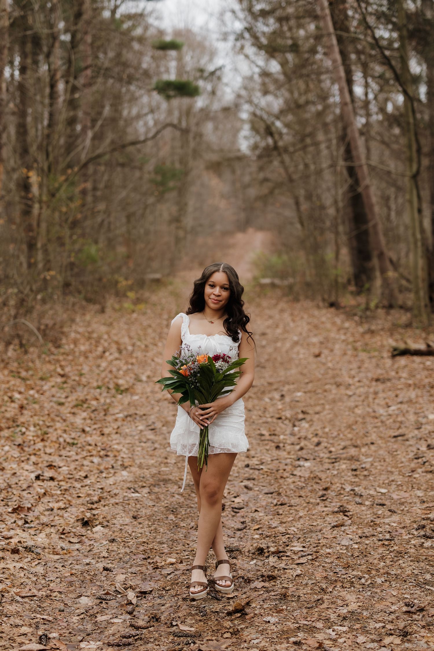 A person in a white dress holds a bouquet while walking on a leaf-covered forest path in autumn.