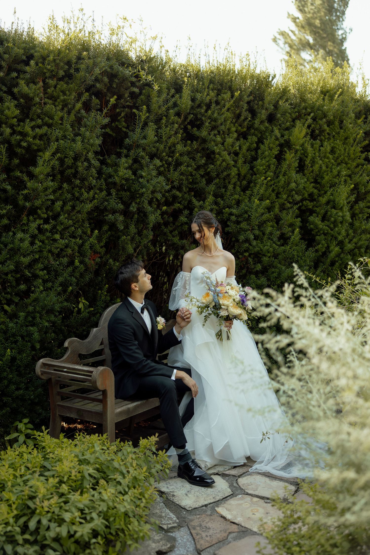 A romantic wedding moment captured on a garden bench surrounded by lush greenery and delicate white flowers.
