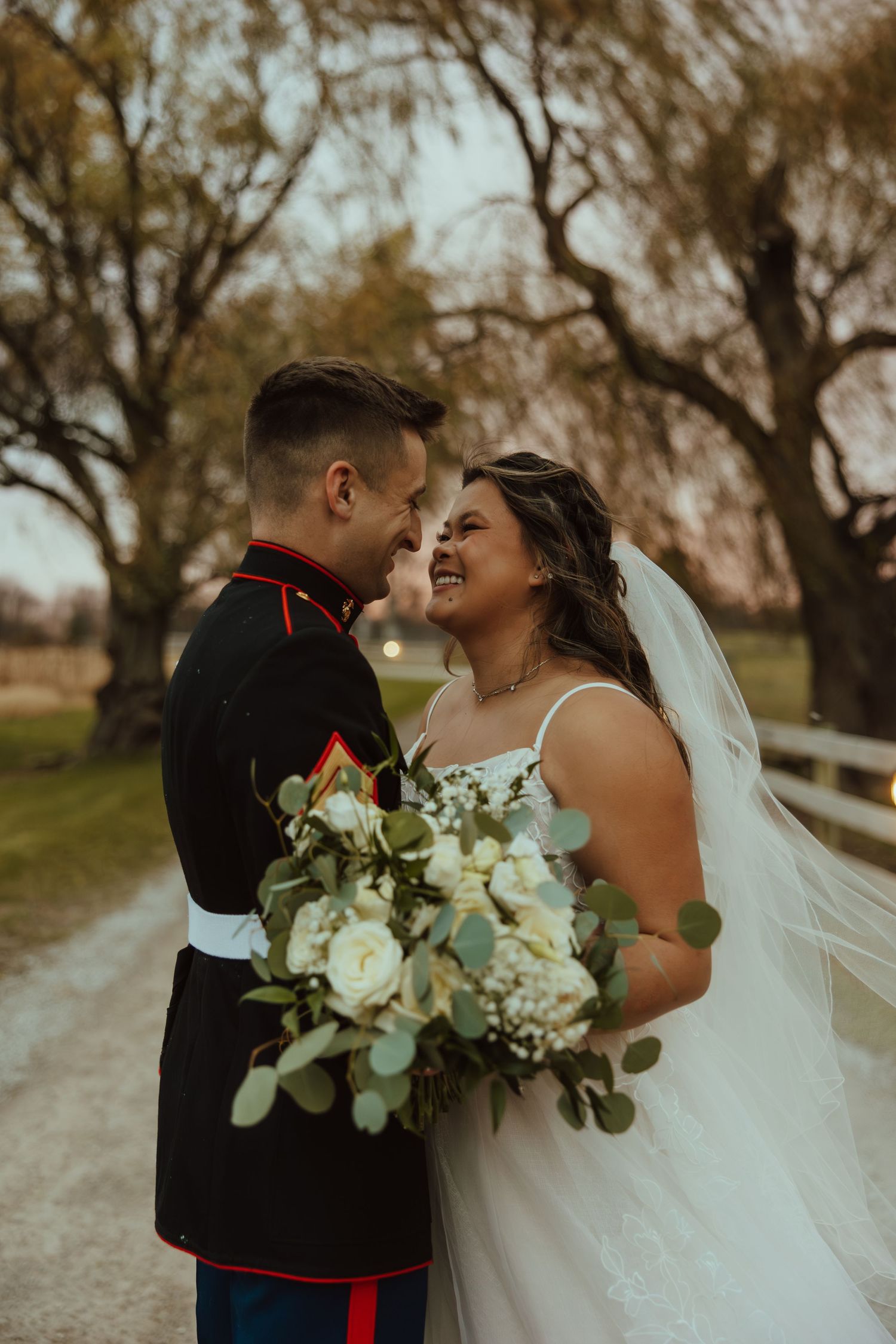 A romantic wedding couple shares intimate moments on a path with trees while holding a white and green floral bouquet.