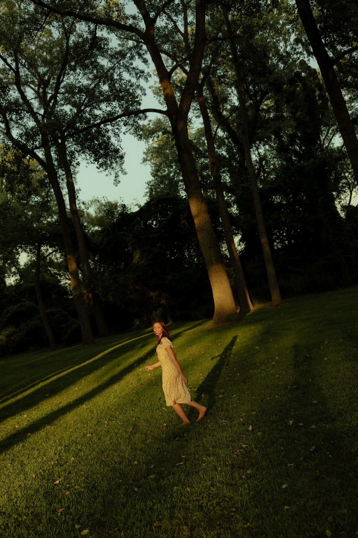 A person runs and leaps across a grassy hillside at sunset with tall trees casting long shadows.