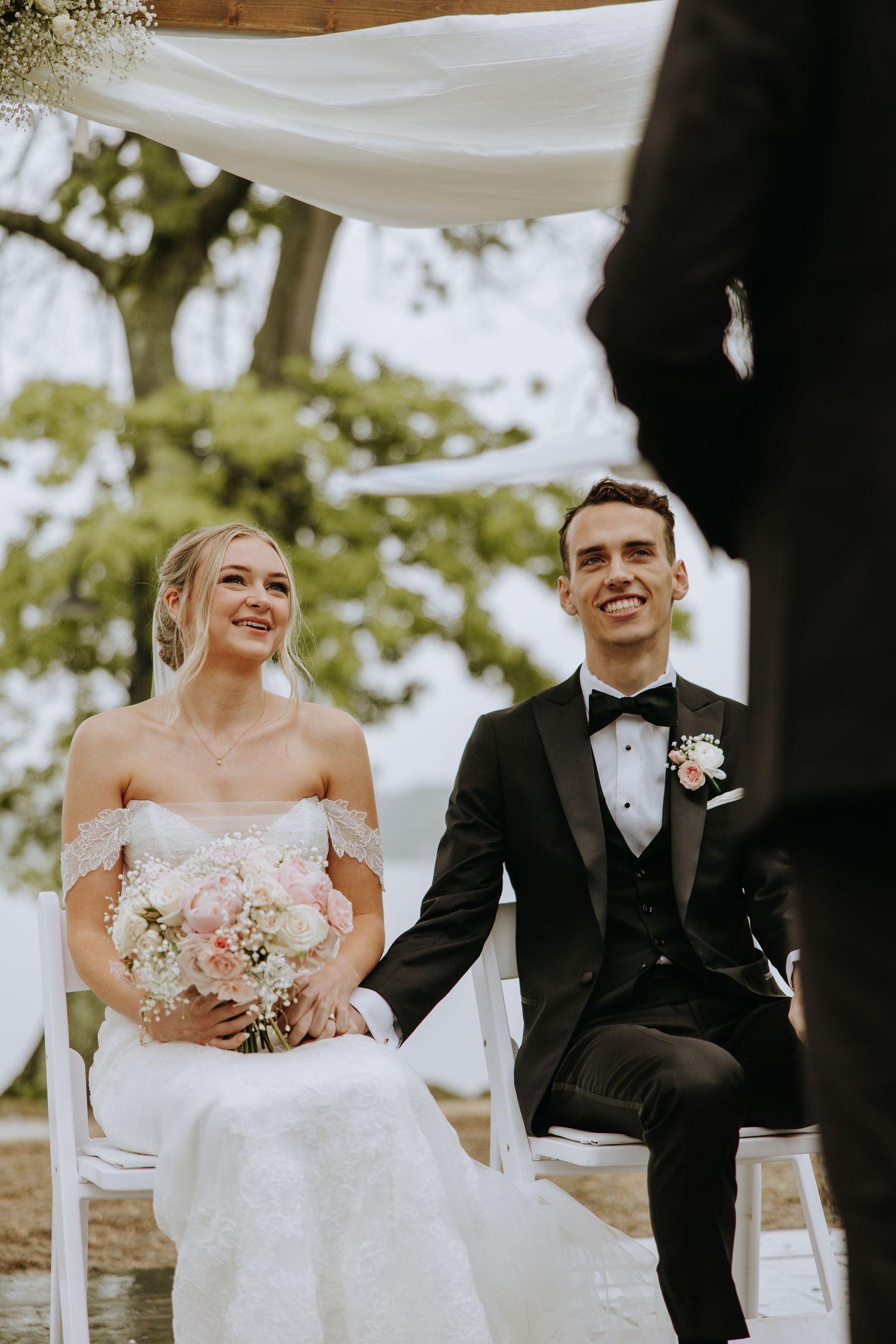 Joyful wedding couple shares laughter during outdoor ceremony under white fabric draping.