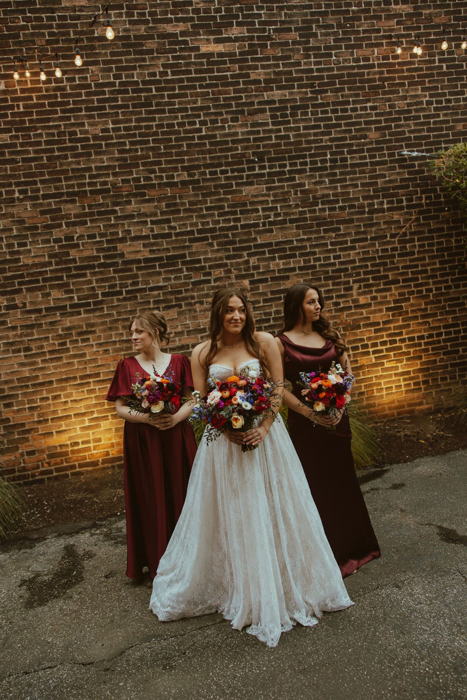 Wedding party members in burgundy dresses and white gown pose against brick wall with floral bouquets at dusk.