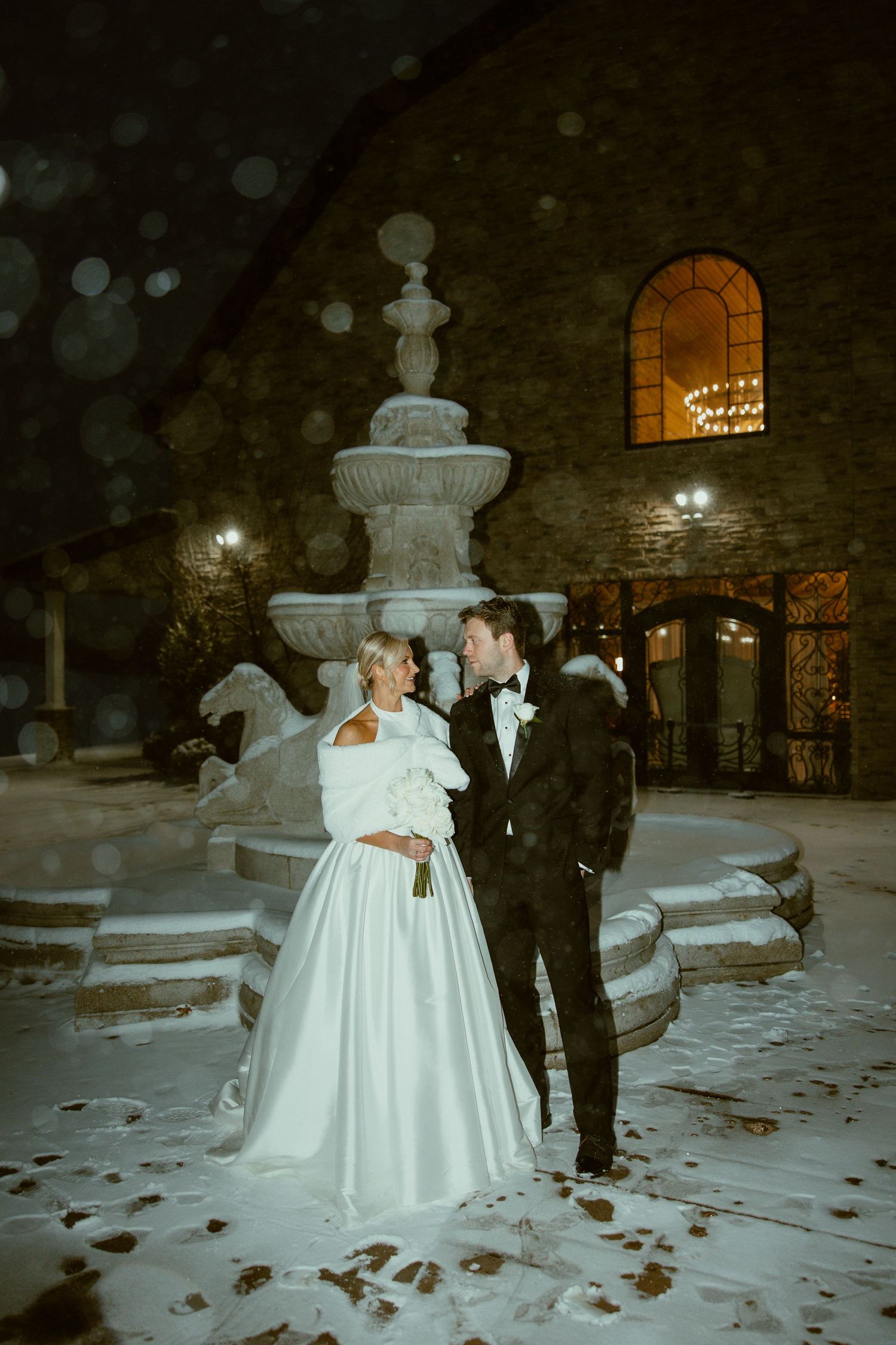 A winter wedding scene with a bride in a white ballgown dancing in the snow near a fountain at night.