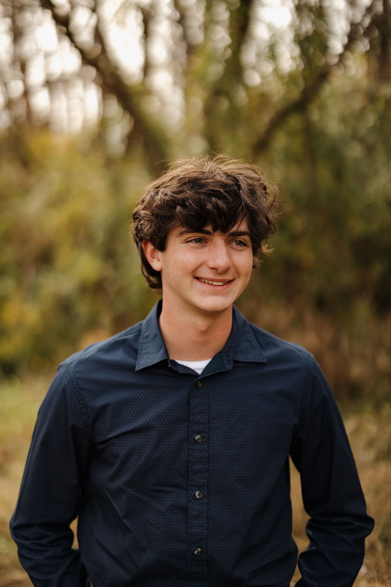A young person in a navy button-down shirt smiles warmly against a blurred forest backdrop.