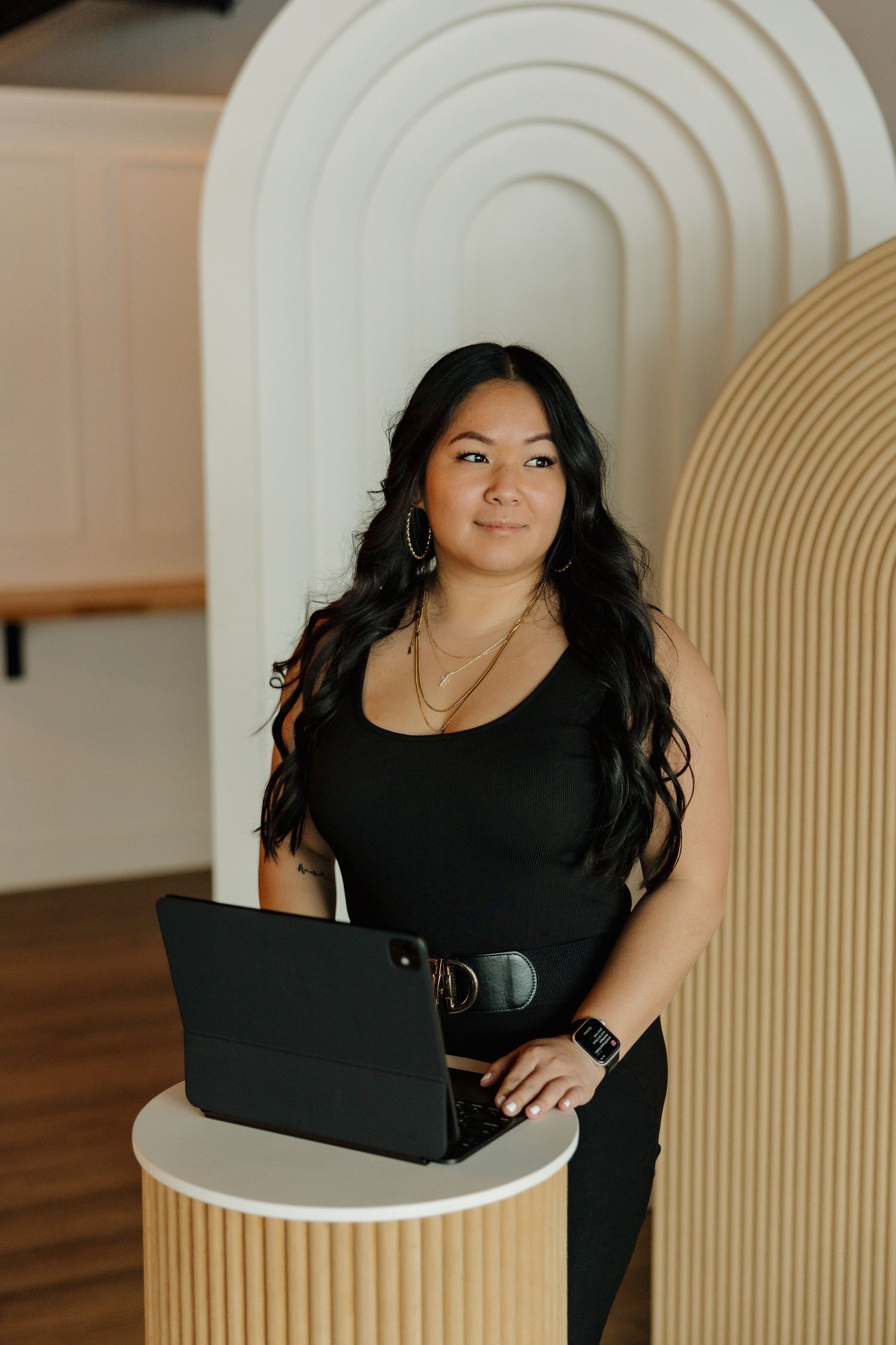 Professional working on laptop against modern white arched wall in stylish black business attire.