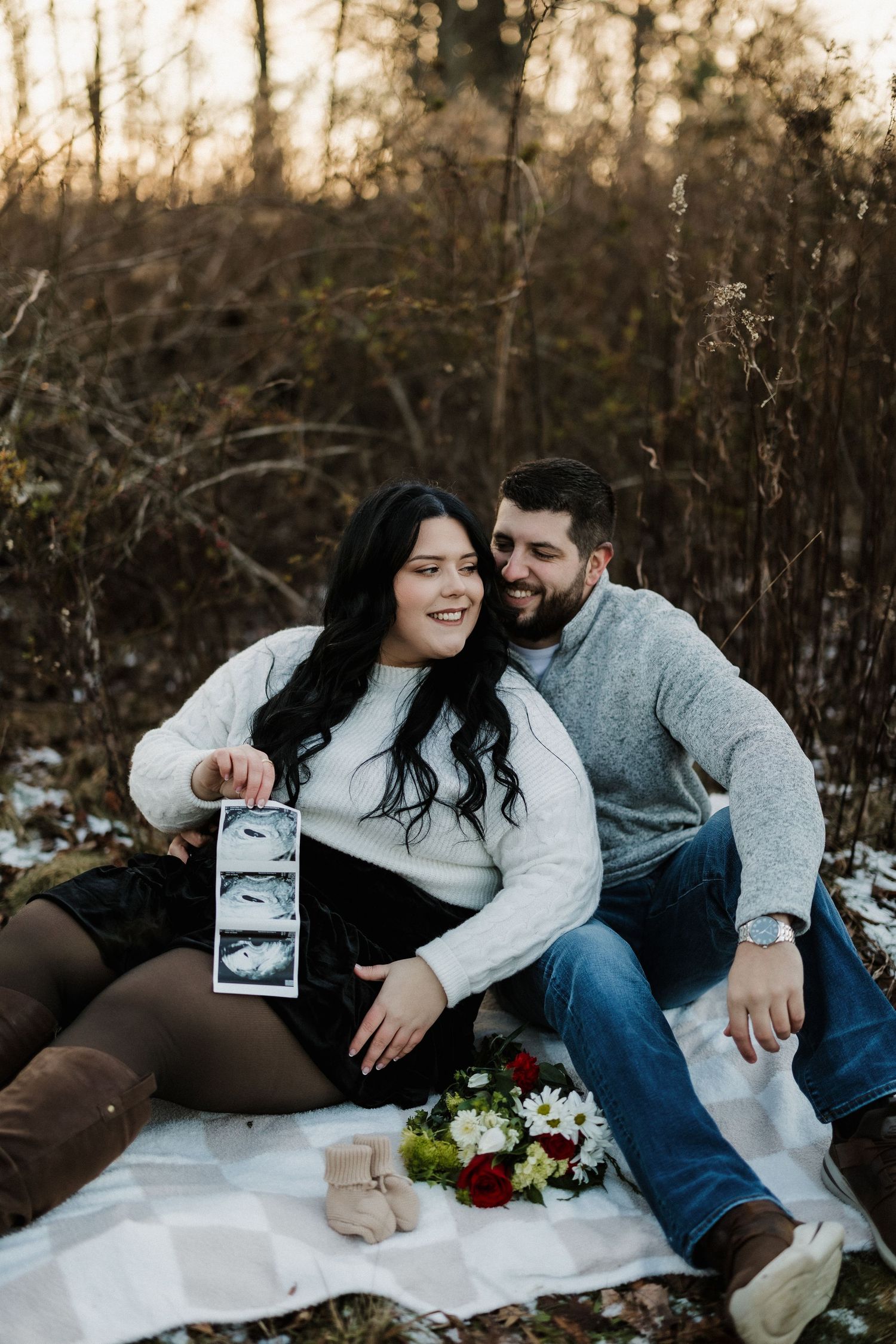 Couple having outdoor photoshoot on blanket with ultrasound photos and flowers.