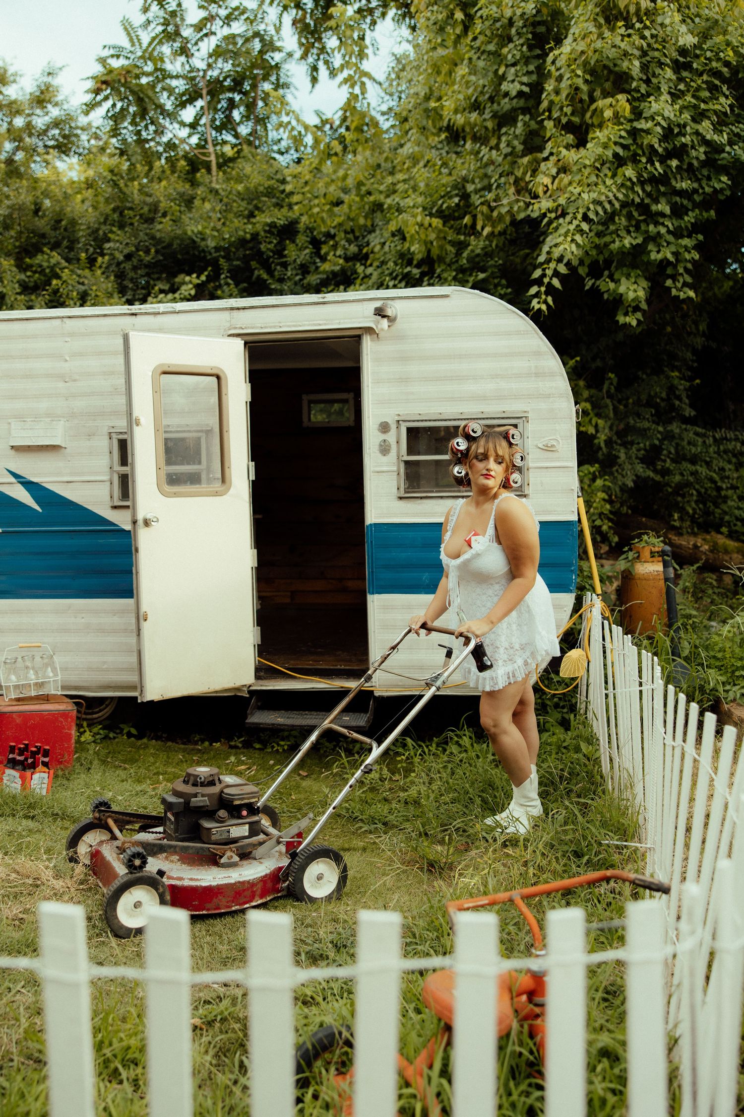 A vintage white and blue camper trailer with a lawnmower in the foreground behind a white picket fence.
