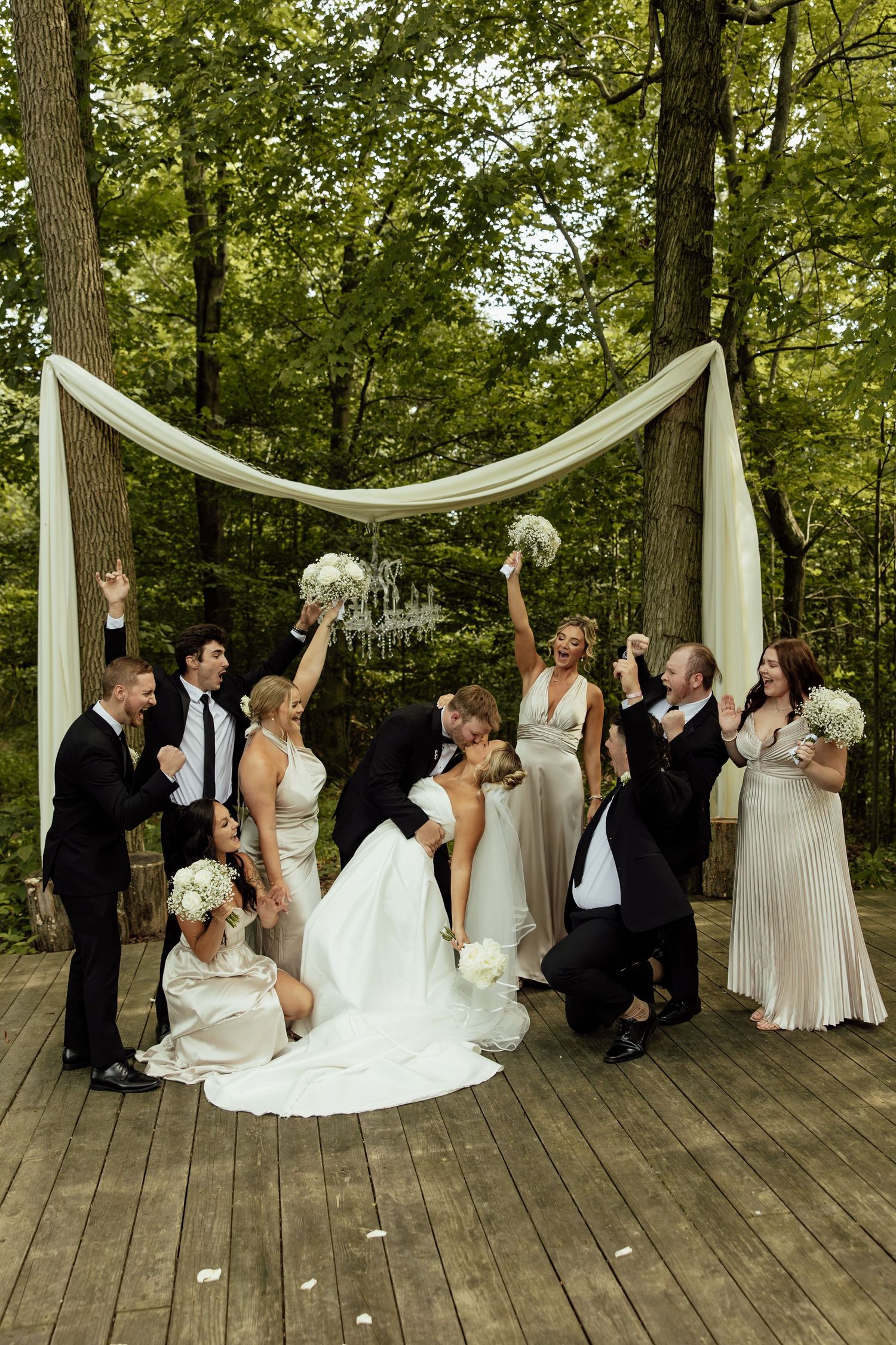 Wedding party celebrates on wooden deck in forest setting with white fabric draped arch and floral bouquets.