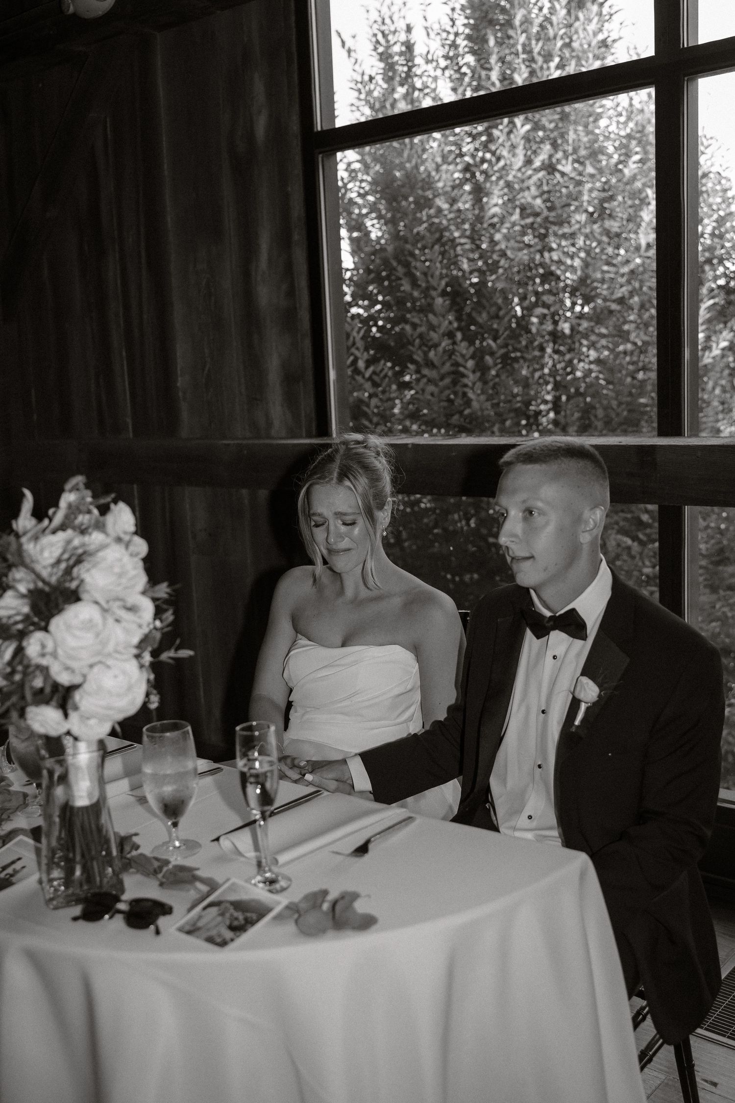 Wedding reception table scene with elegant white tablecloth and floral centerpiece by large windows.