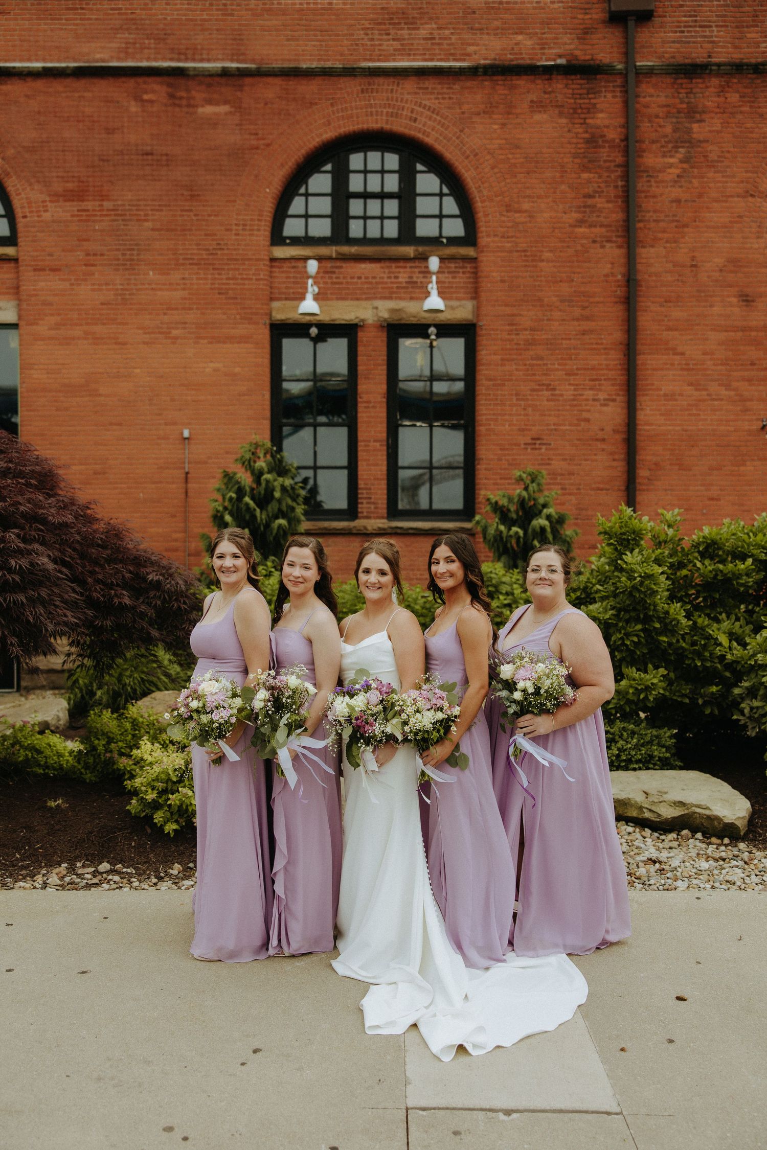 Wedding party poses outside a brick building with bridesmaids in lavender dresses alongside a bride in white.