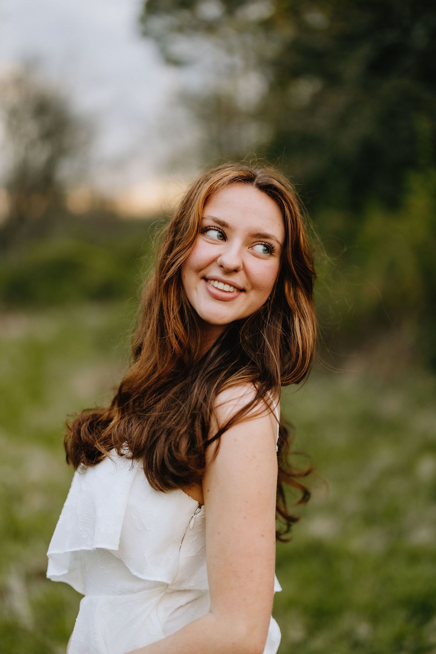 A graduate in a white ruffled dress poses outdoors in natural lighting.