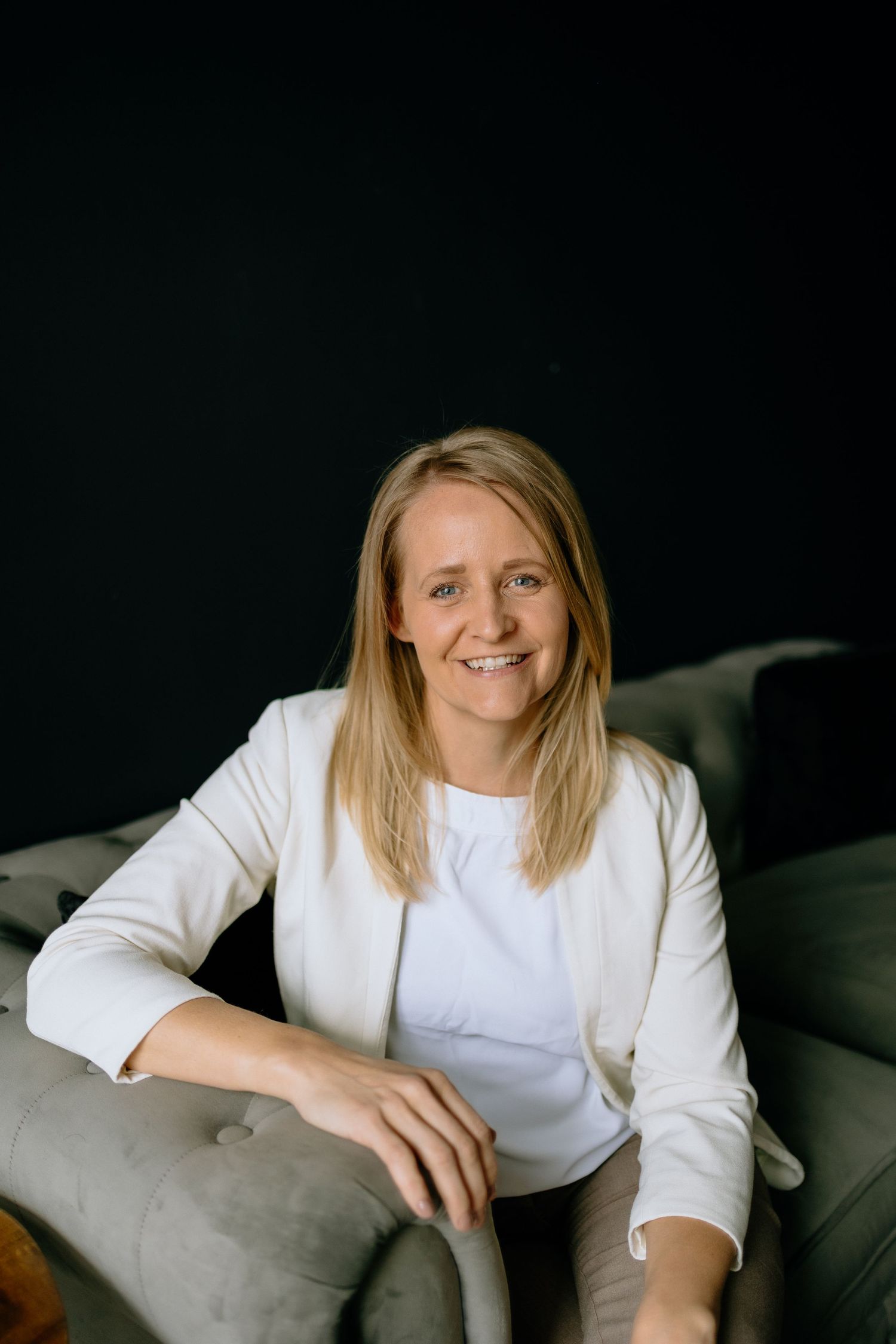 Professional portrait of person in white top sitting on gray couch against dark background.